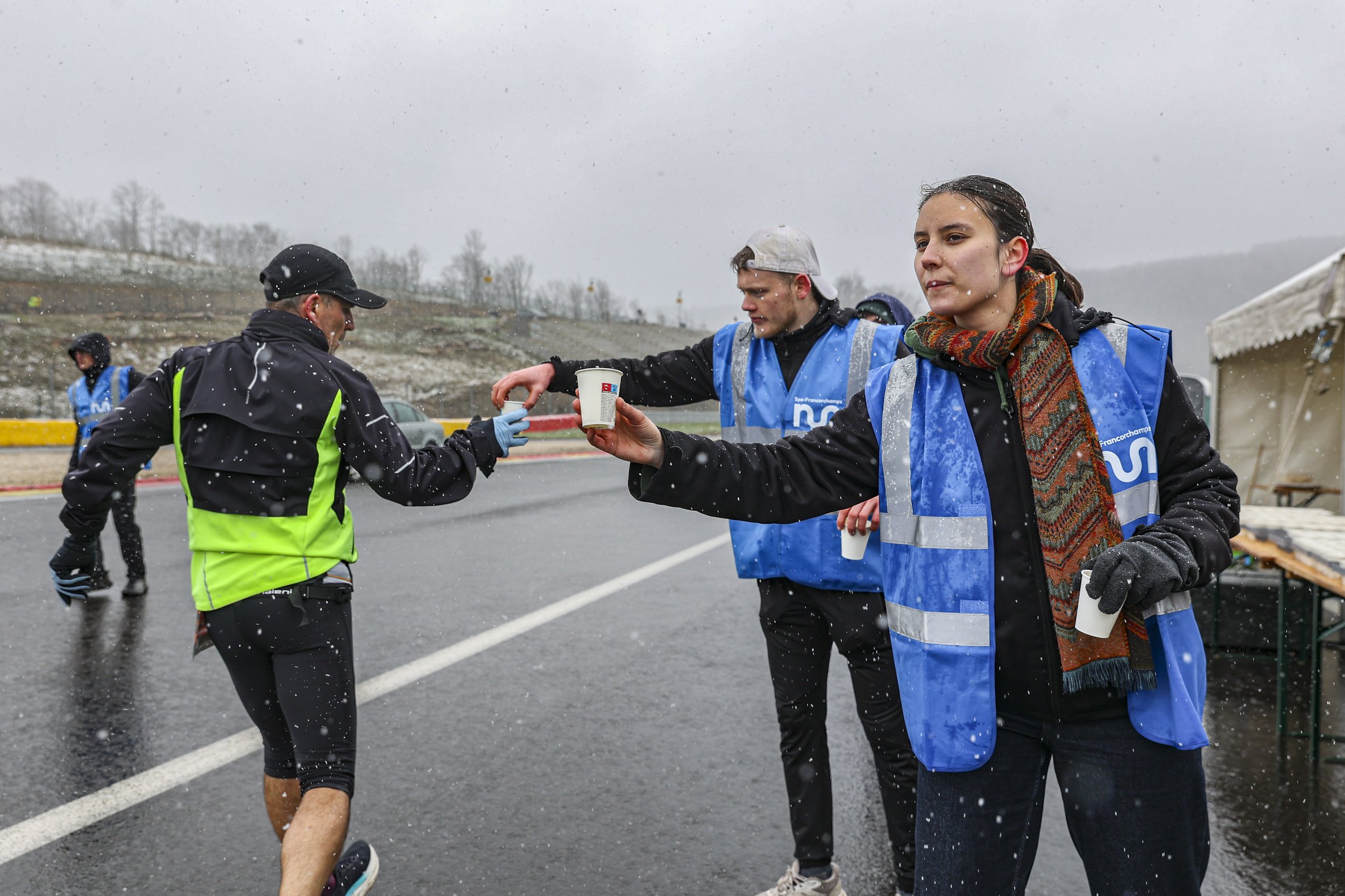 A person in athletic gear runs through rain while two volunteers in blue vests hand out cups on a wet road during a race or marathon.