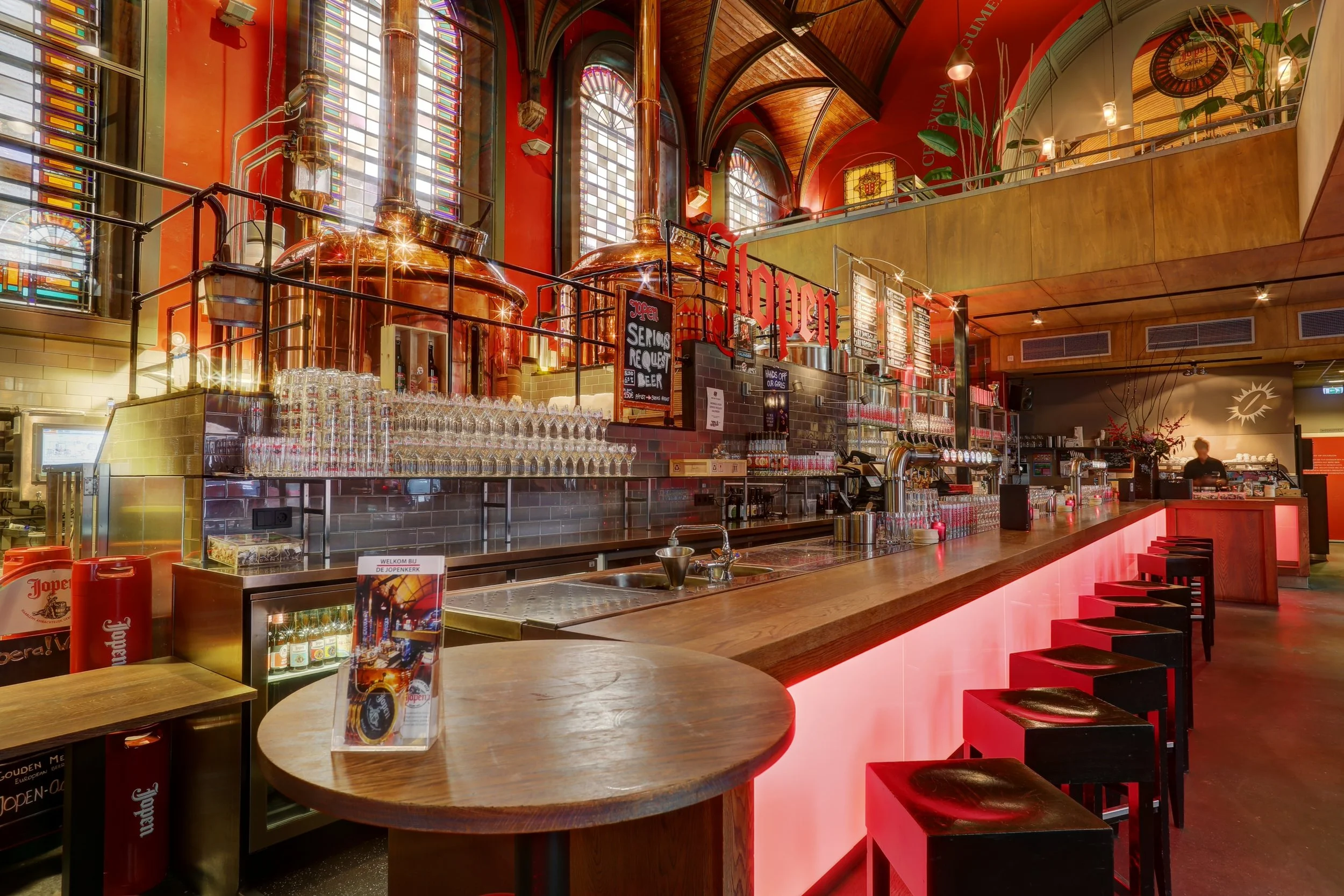Interior of a bar or restaurant with large stained glass windows, copper brewing equipment, a wooden counter with red stools, and a chalkboard menu.