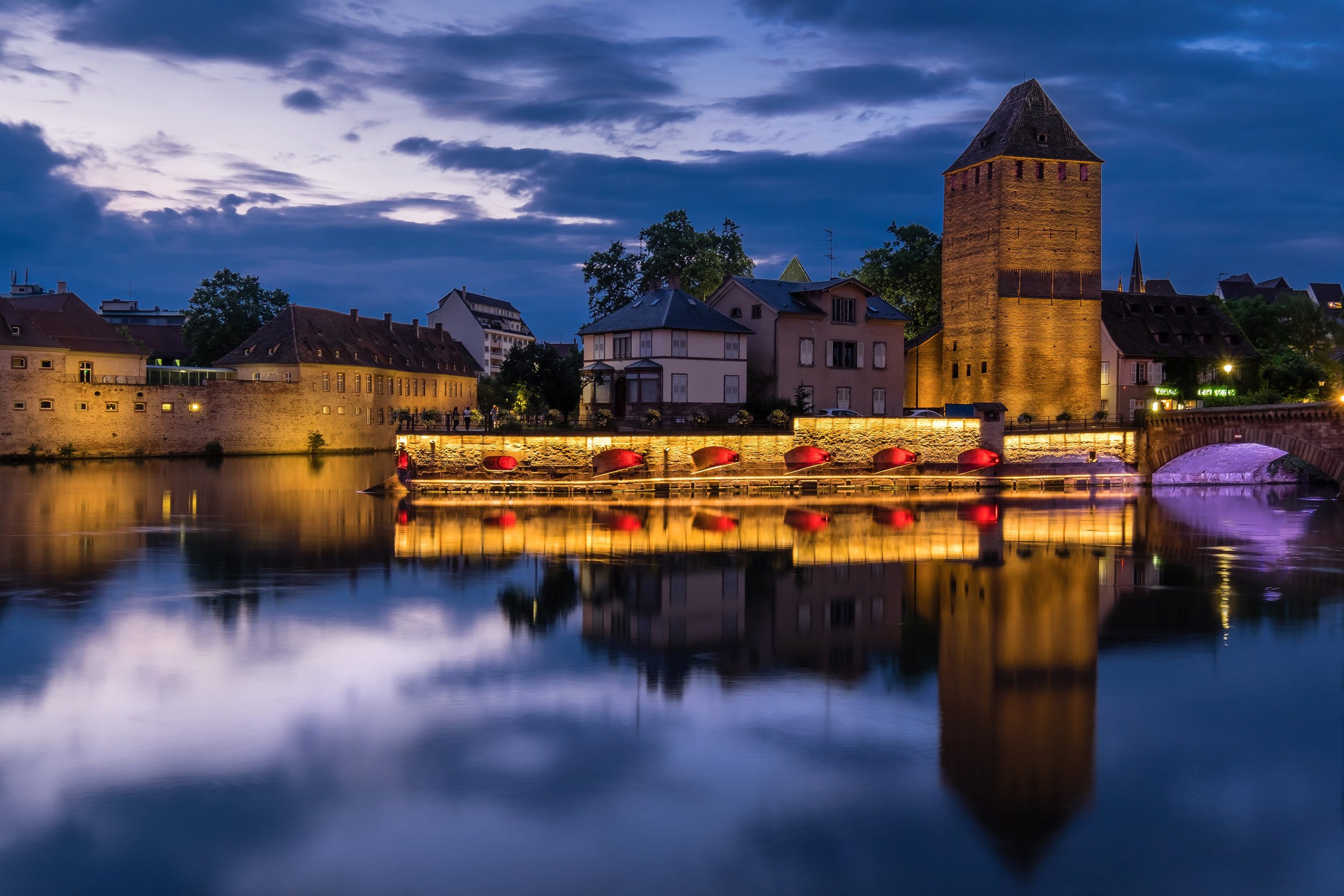 Nighttime view of a historic European town with illuminated buildings, a prominent tall brick tower, and a bridge over a calm river, with a cloudy sky overhead.