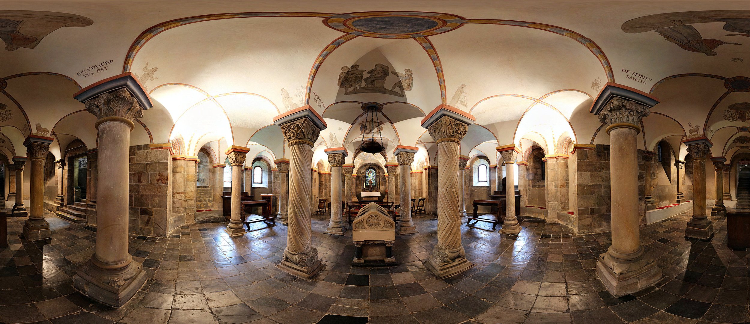 Interior view of a historic church with stone columns, arched ceiling, and religious artwork, featuring a central altar space with seating.