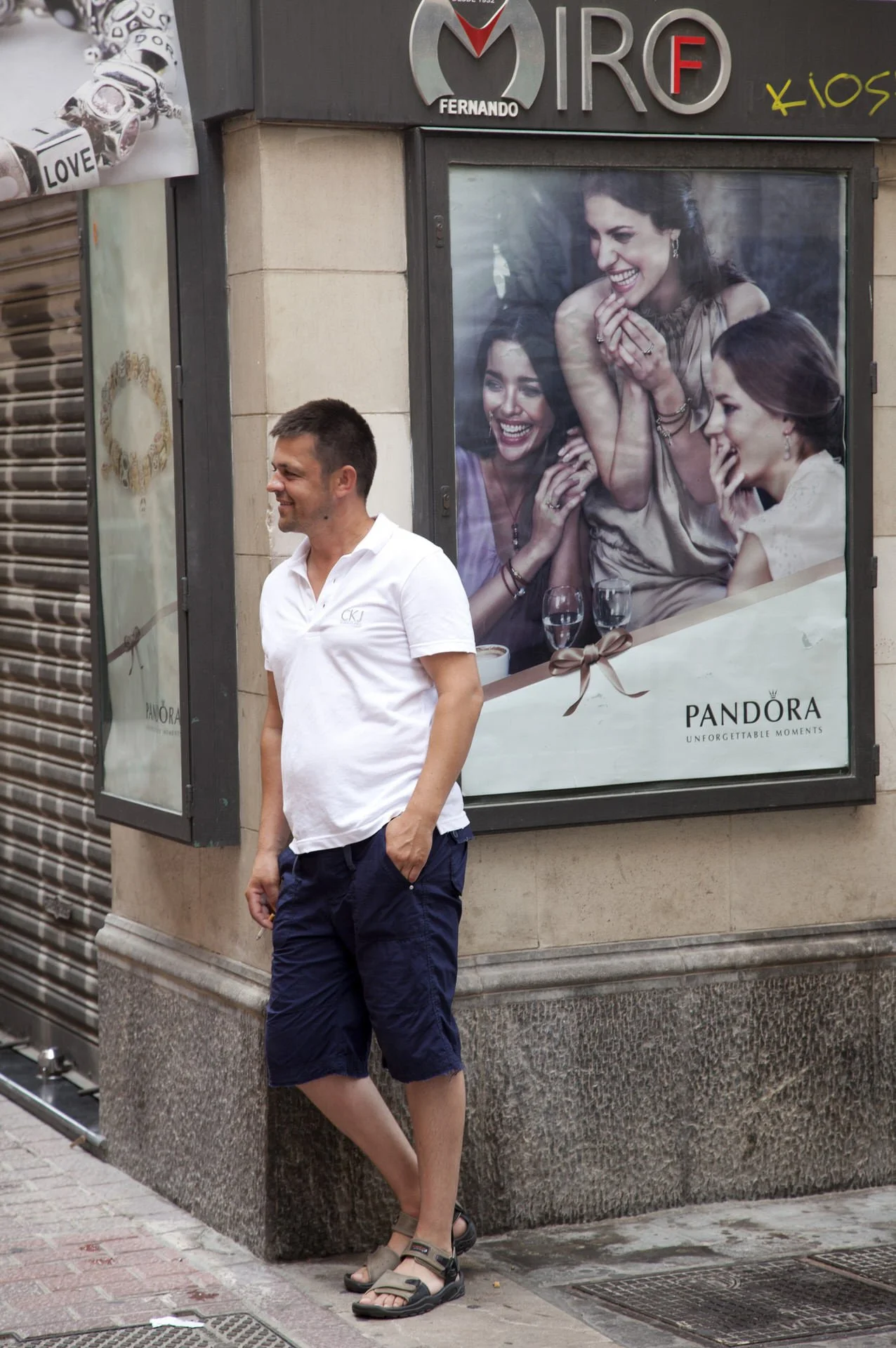 A young man in a white polo shirt and navy shorts standing on a city sidewalk in front of a large window displaying an advertisement for Pandora jewelry. The advertisement shows three women laughing and enjoying each other's company. The man is smili