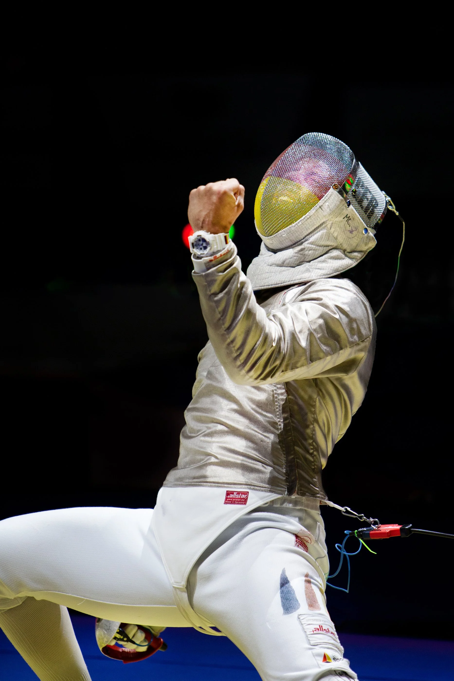 An Olympic fencer in white uniform and protective mask celebrating with a fist pump.
