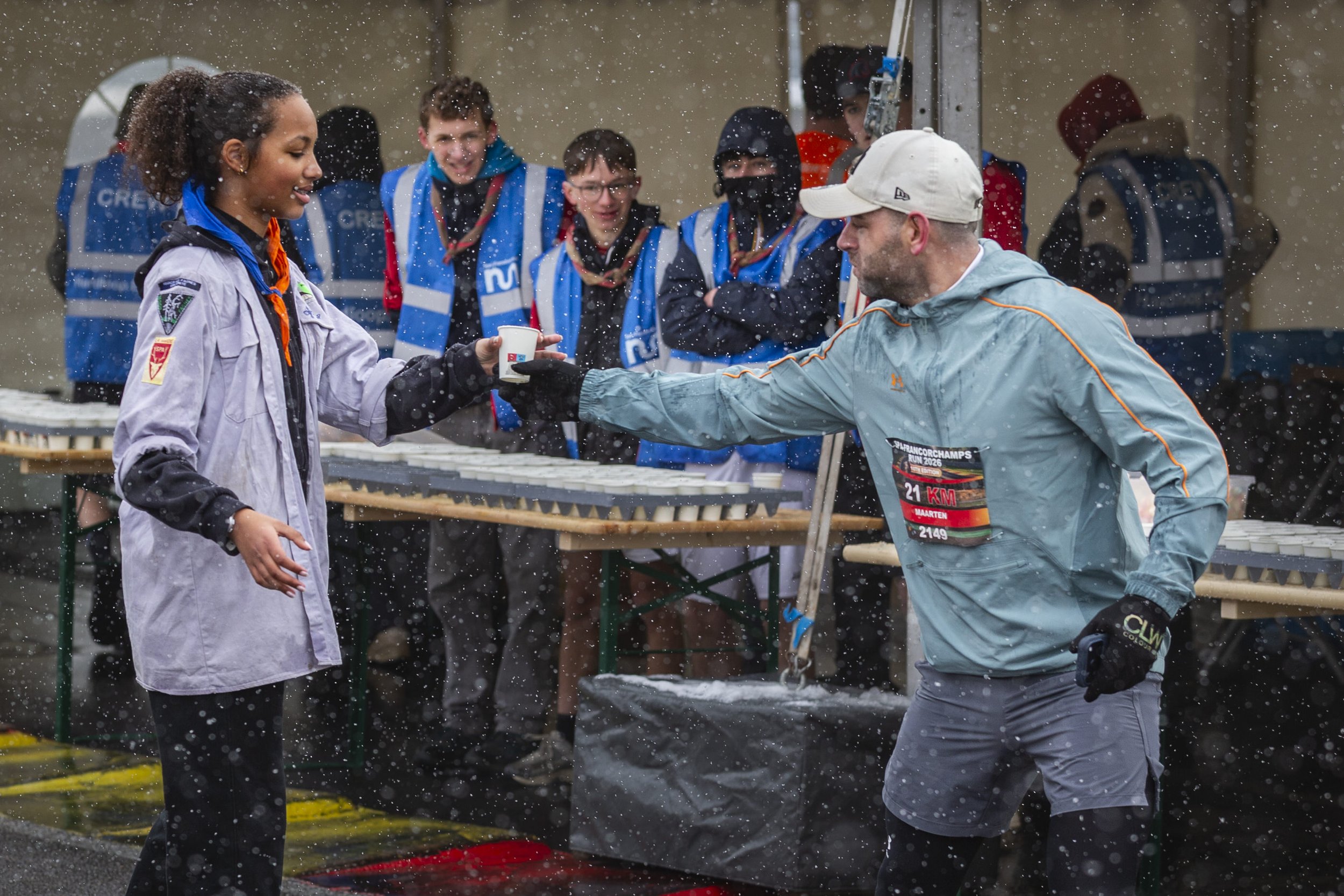 A woman in a white jacket receiving a beverage from a man in a gray jacket and white cap at an outdoor event on a rainy day, with a group of people watching in the background.