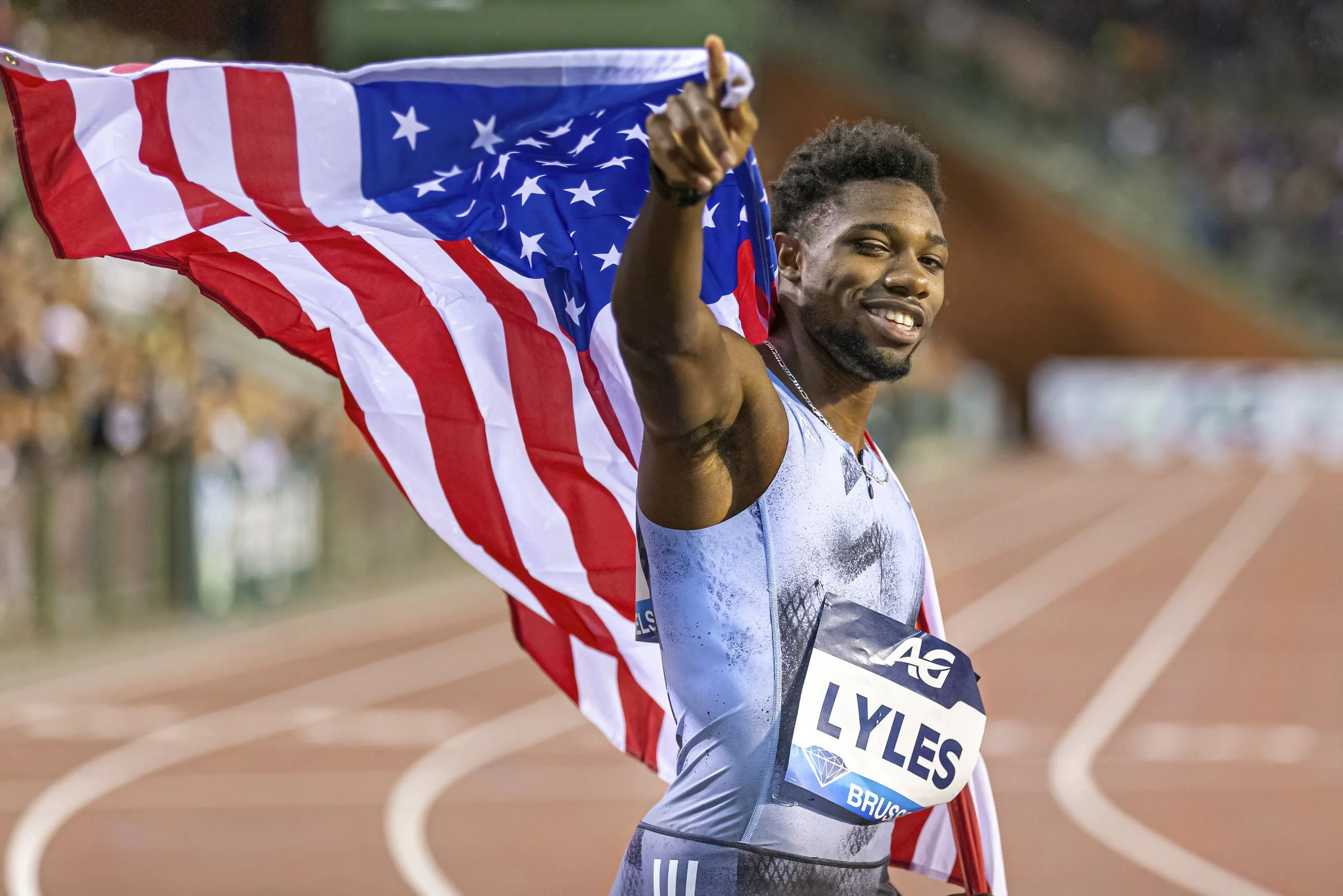 A male athlete holding an American flag and smiling on a track field, wearing a silver athletic suit with a bib number that reads Lyles.