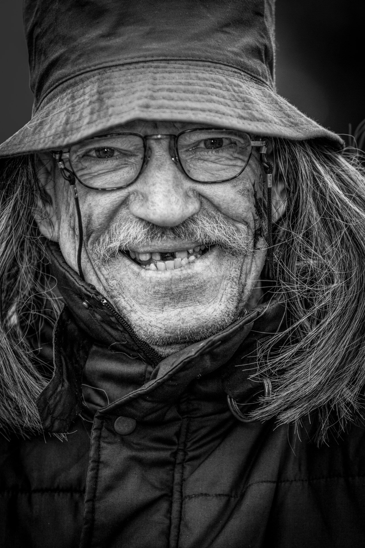 Close-up black and white photo of an older man with long hair and a beard wearing glasses, a hat, and a winter coat.