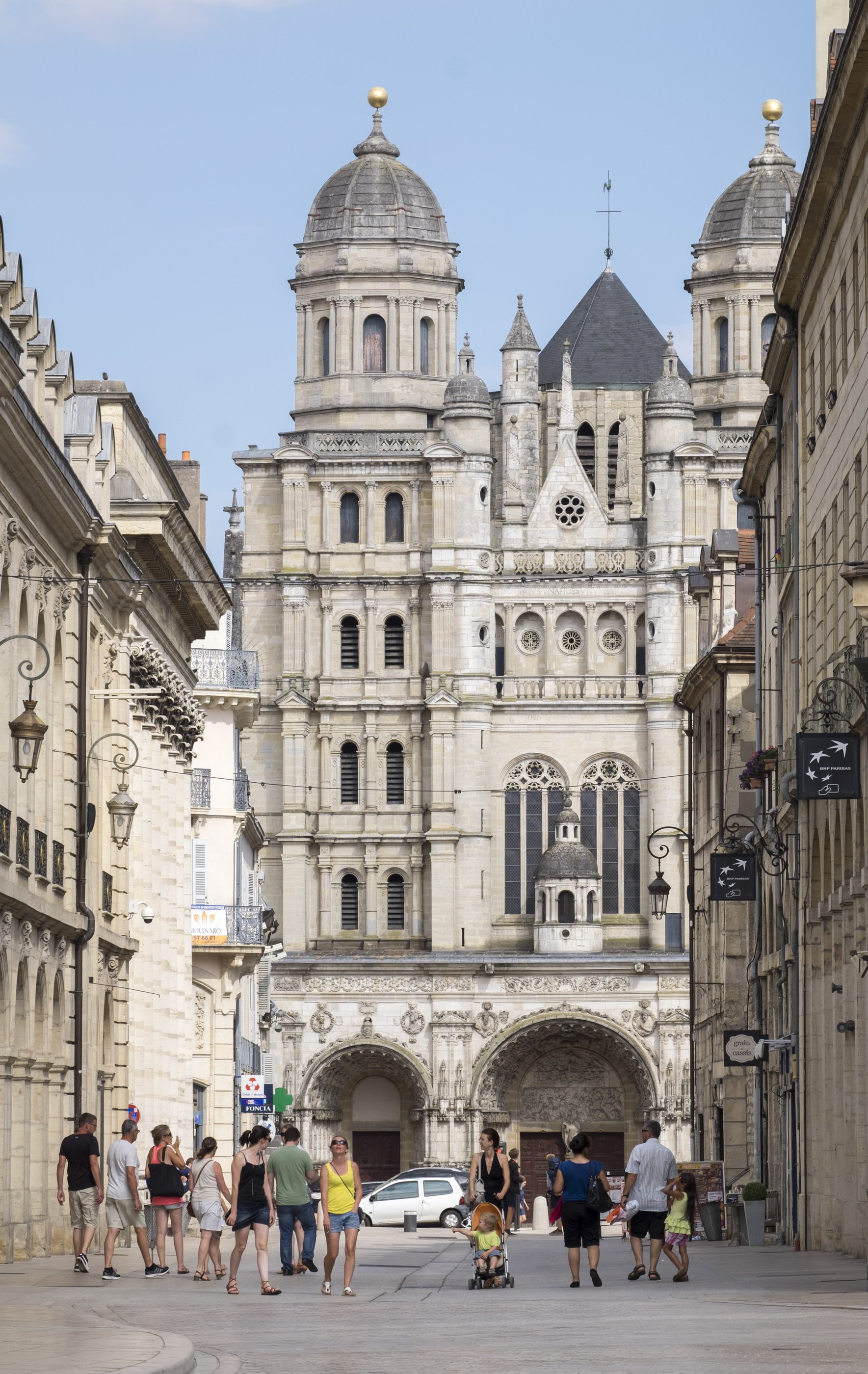 A busy street scene in front of a large historic church with twin towers and detailed stone architecture. People are walking on the street, some with children, and a car is parked nearby. The sky is blue with some clouds.