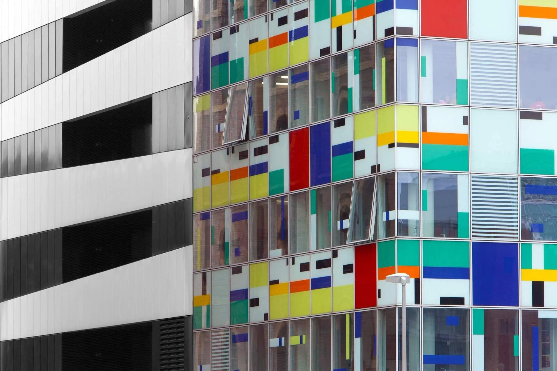 A modern building with a black and white striped facade on the left and a colorful, geometric window design on the right. The windows feature various small, brightly colored rectangles and squares in yellow, orange, blue, green, red, and black.