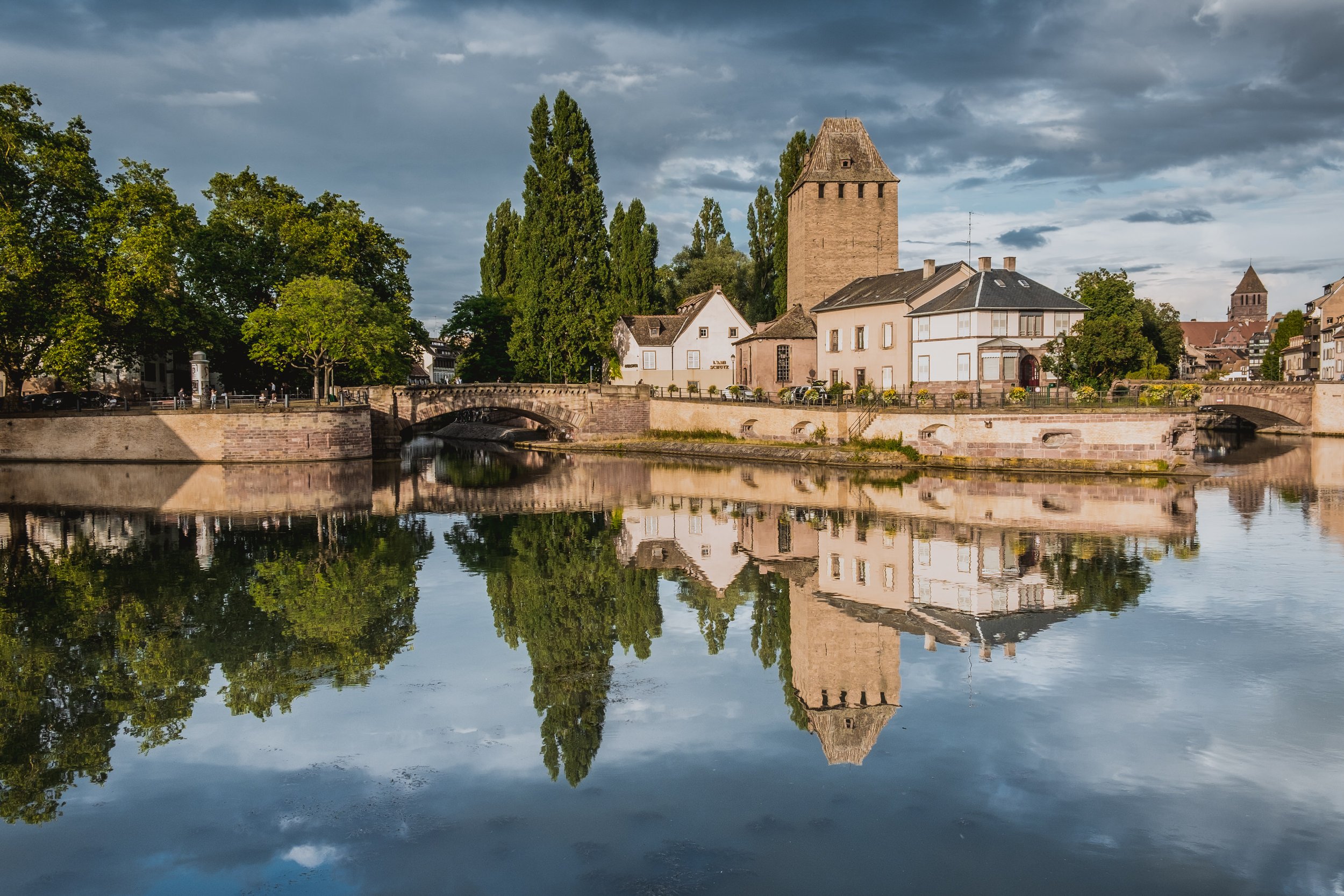 A scenic view of a historic tower and buildings along a river, with reflections in the water and a partly cloudy sky.