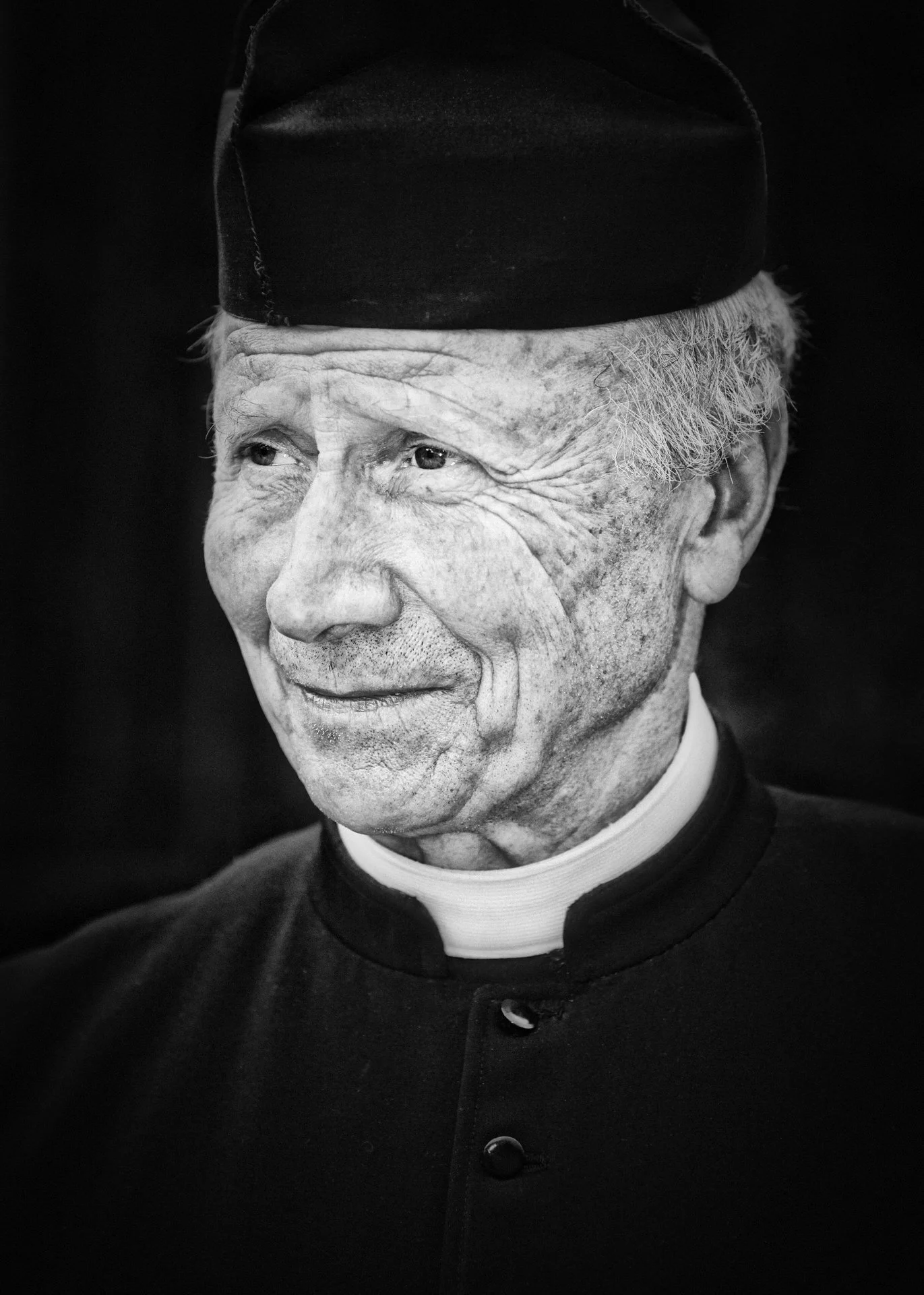 Black and white photograph of an elderly man with wrinkles smiling, wearing a clergy outfit with a clerical collar and a black hat.
