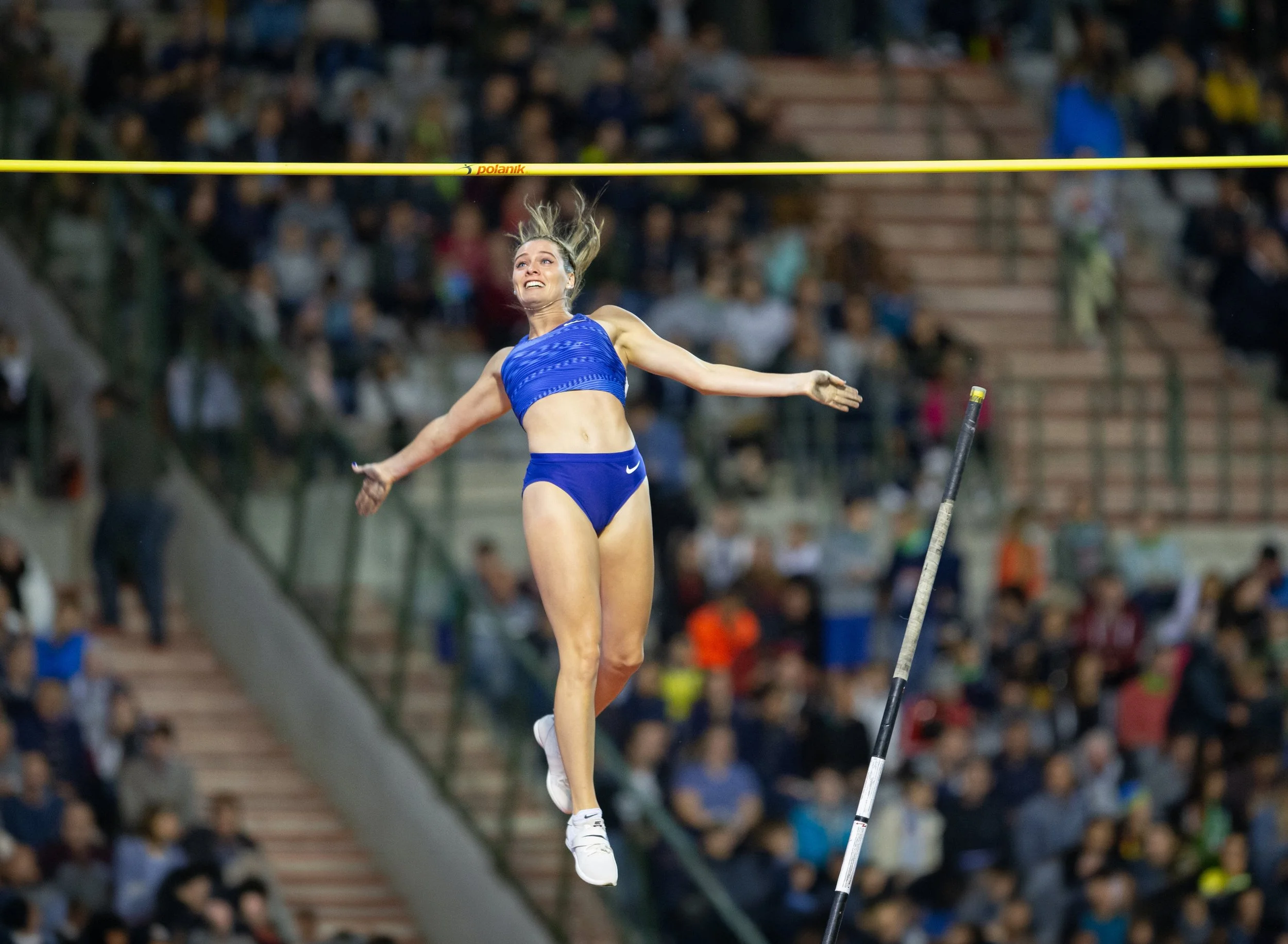 A female tennis player in a blue sports outfit is mid-air, attempting to hit a tennis ball with her racket during a match in an indoor stadium crowded with spectators.