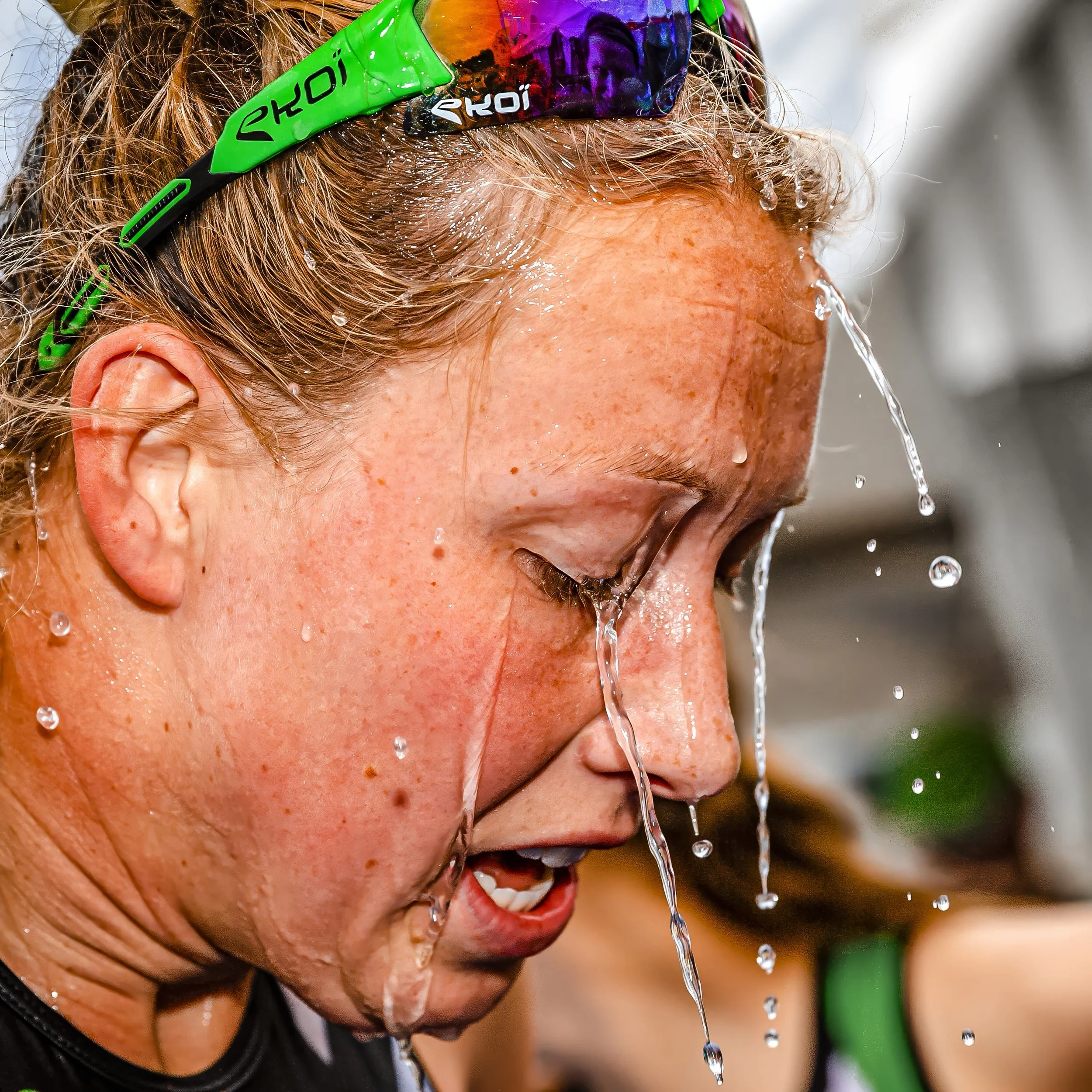 A person with wet hair is wearing sunglasses and appears to be smiling while water is cascading down their face.
