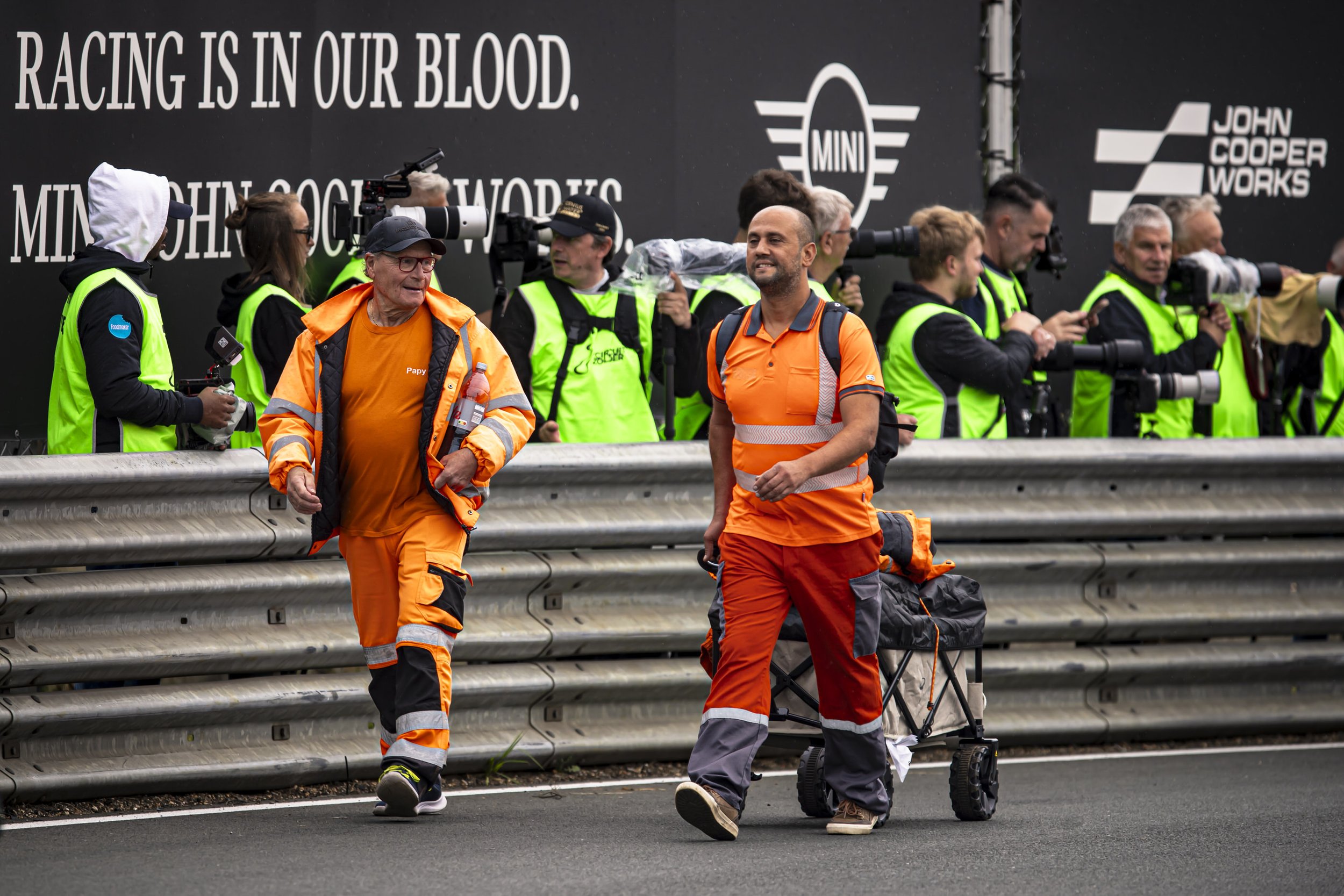 Two men in orange and black work uniforms walking along a road with a metal guardrail, while a group of photographers and videographers with cameras and zoom lenses stand behind the guardrail, taking photos of them. There is a large black banner in t