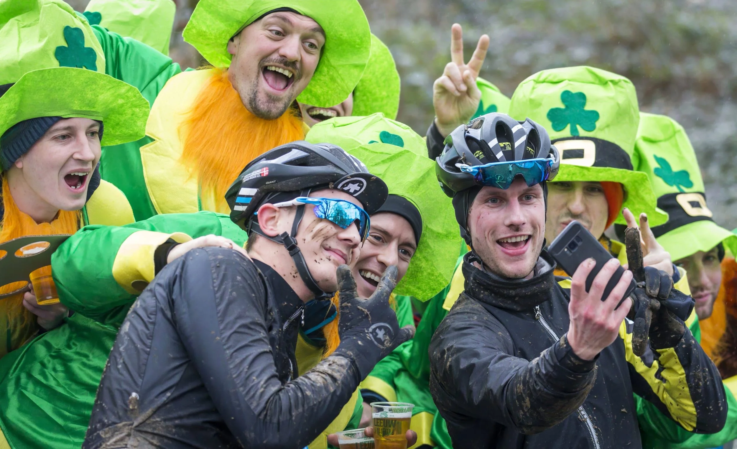 A group of people dressed in green shamrock-themed costumes and helmets, celebrating outdoors, with some holding drinks and making cheerful gestures.