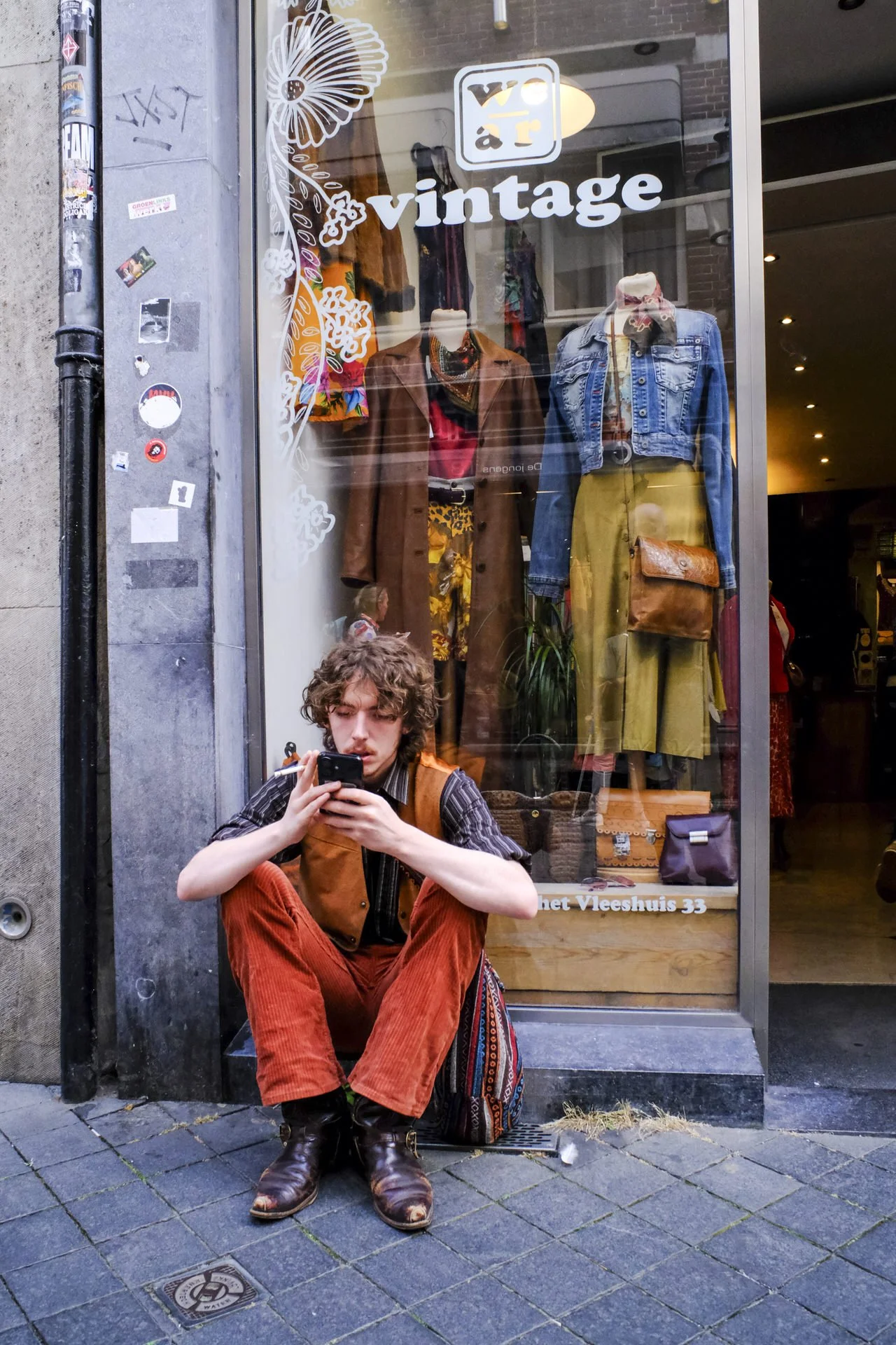 A young man with curly hair sitting on the sidewalk, wearing a striped shirt, brown vest, red pants, and brown boots, looking at his phone. Behind him is a clothing store window display with mannequins dressed in vintage clothes, a sign reading "vint