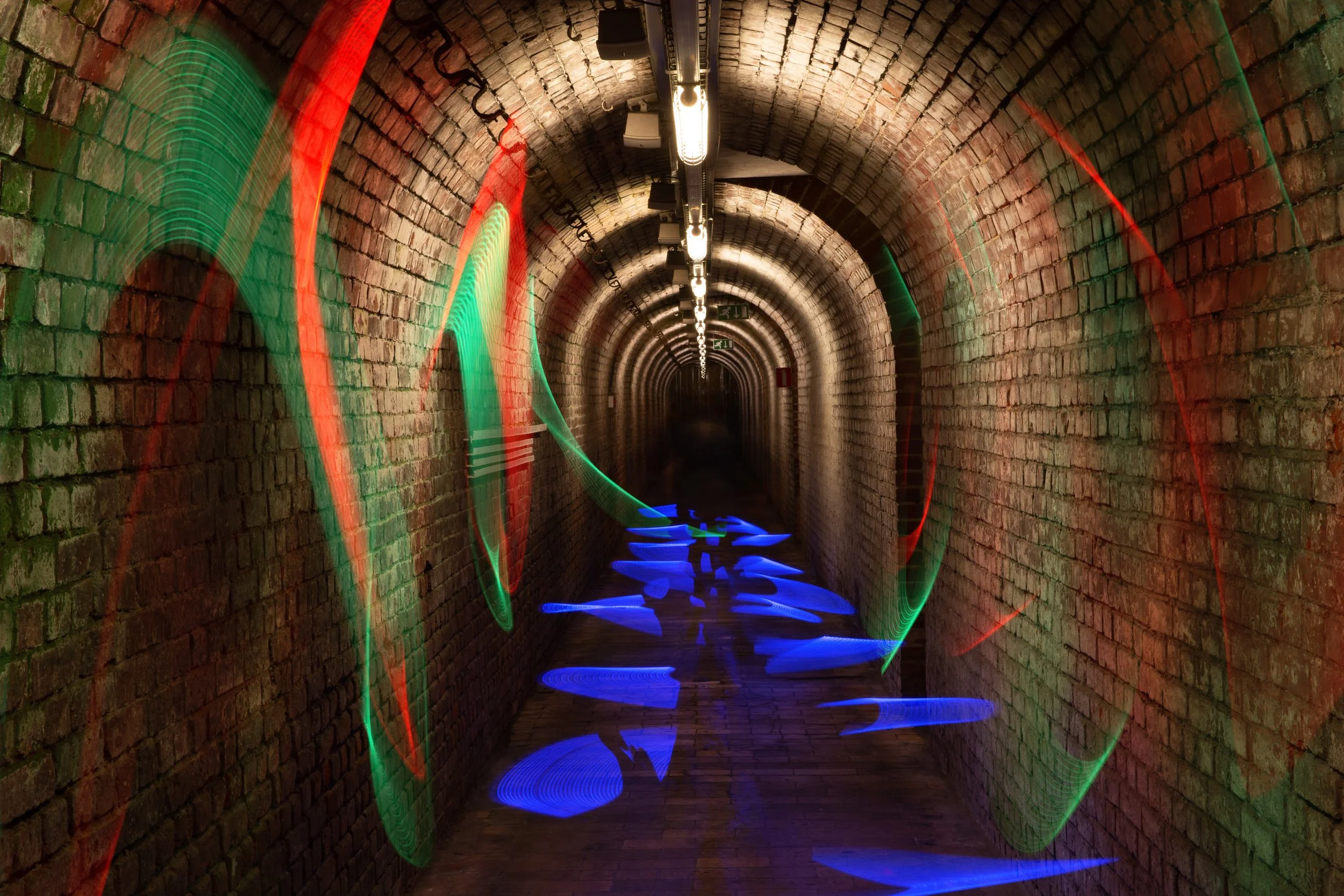 Underground brick tunnel with fluorescent colored light trails and reflections, illuminated by overhead lights.