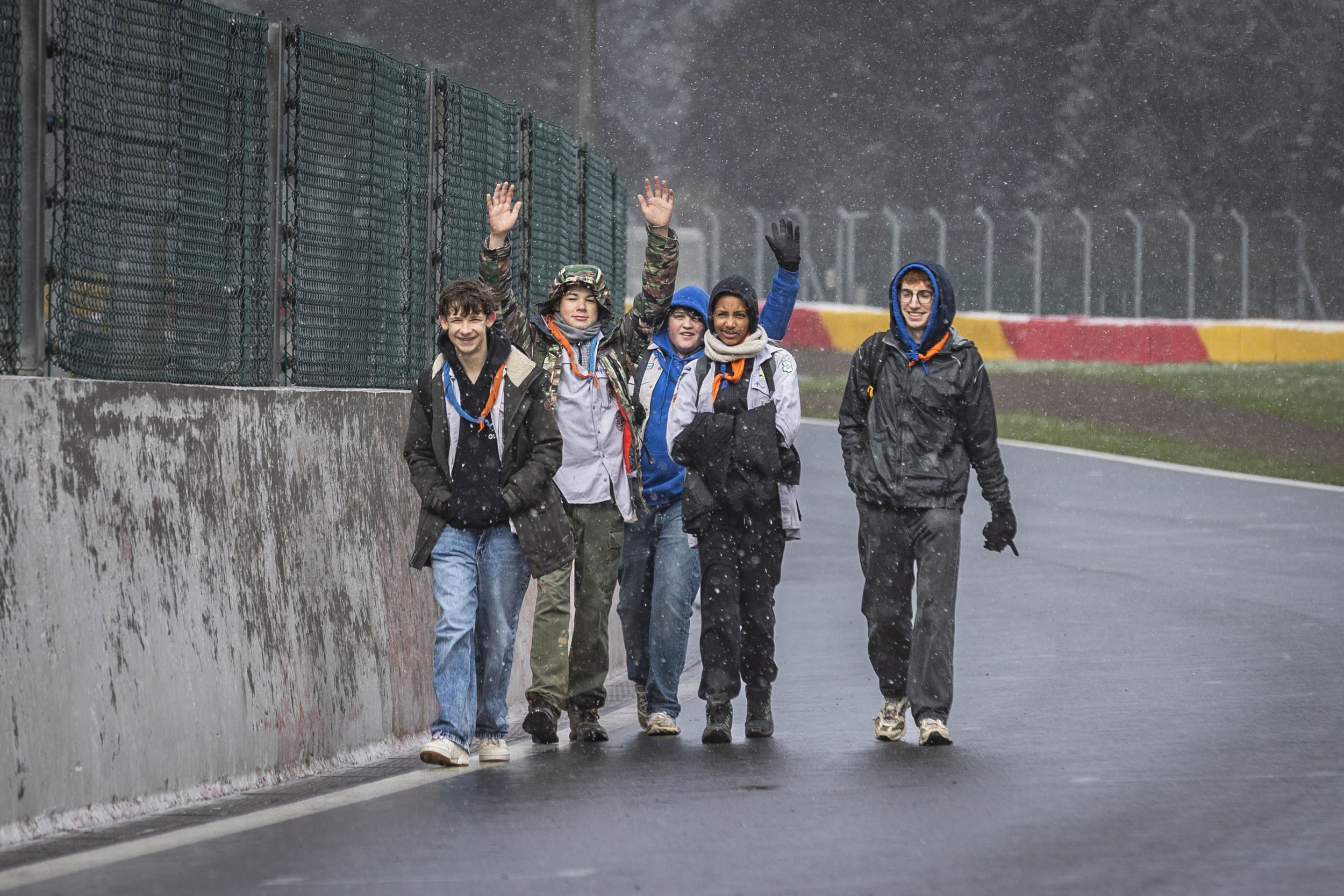 Group of young people walking along a rainy track next to a barrier fence, with some raising their hands and smiling, wearing jackets and rain gear.