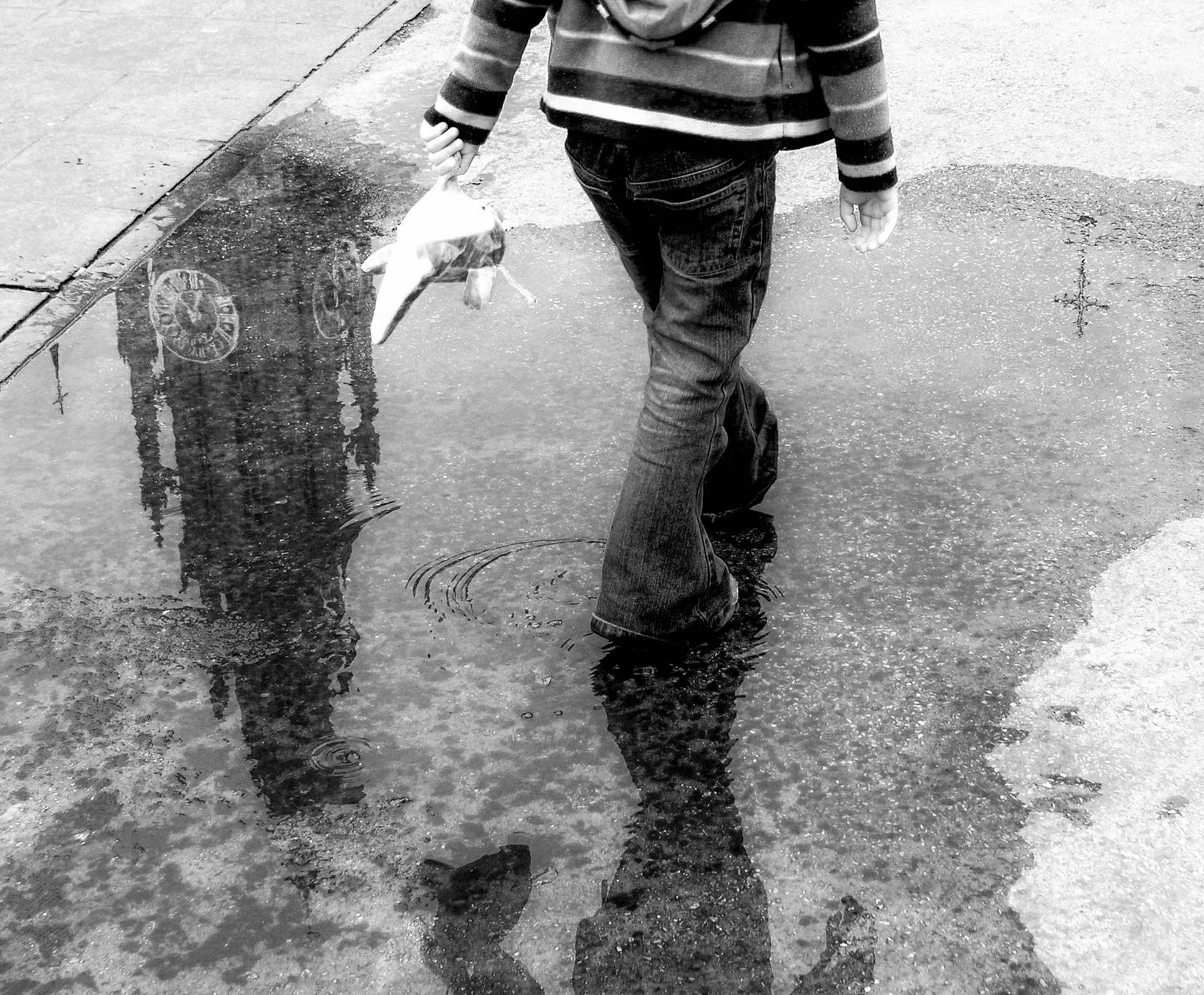 A child holding a dead fish by the water's edge on a city sidewalk in black and white.