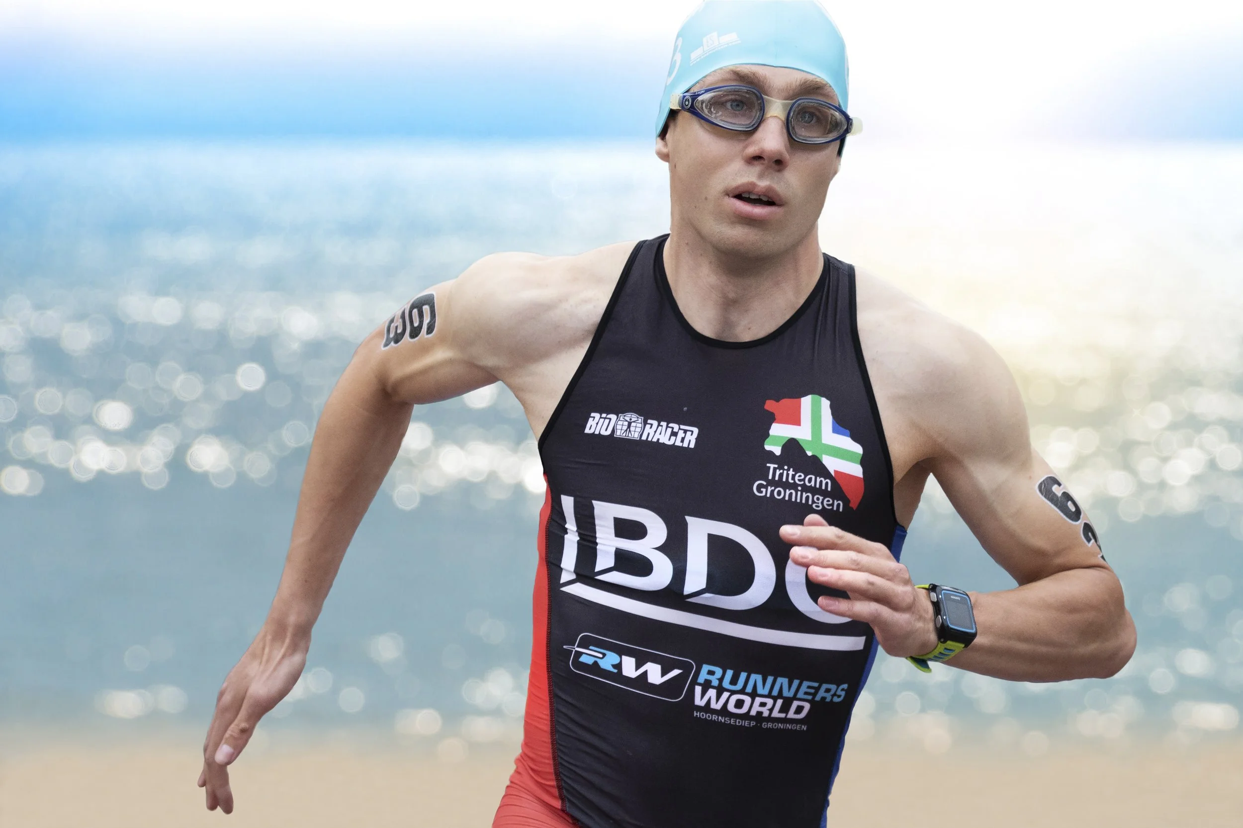 A male triathlete running on a beach, wearing a blue swim cap, goggles, and a black, red, and white racing suit with various logos, including one of a map of the Netherlands and the word 'BID' on it.