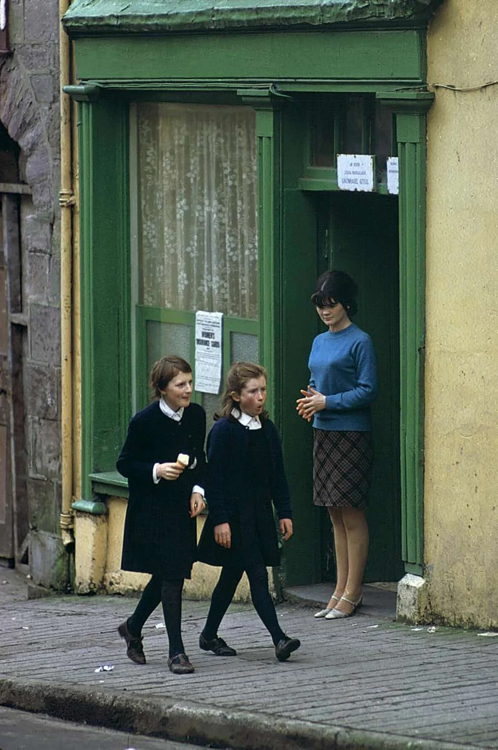 A woman standing in a doorway watching two young girls walk by on the sidewalk, one of whom is eating an ice cream cone.