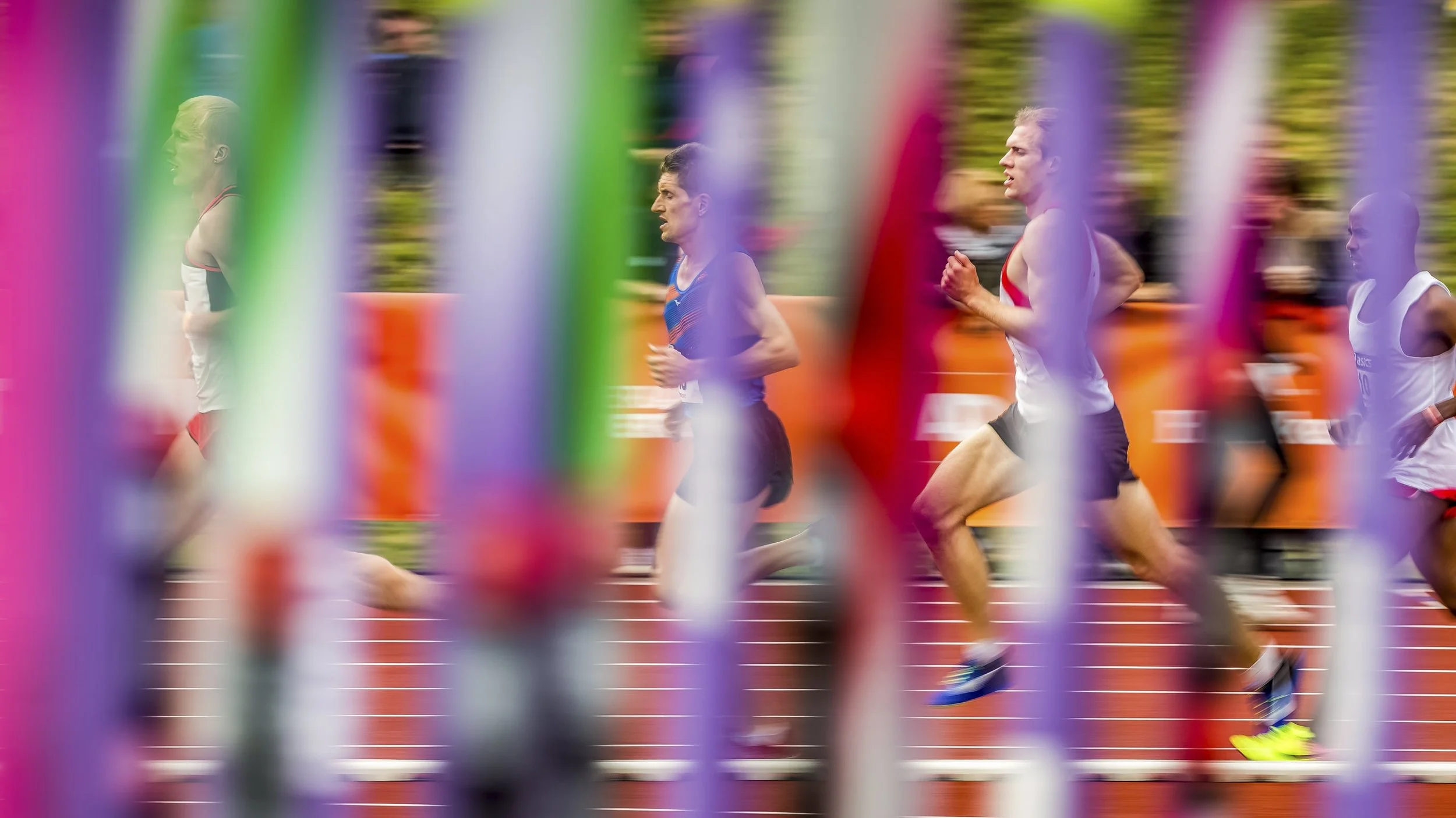 A group of marathon runners competing on a track field, with colorful barriers or flags in the foreground creating a blurred pattern.