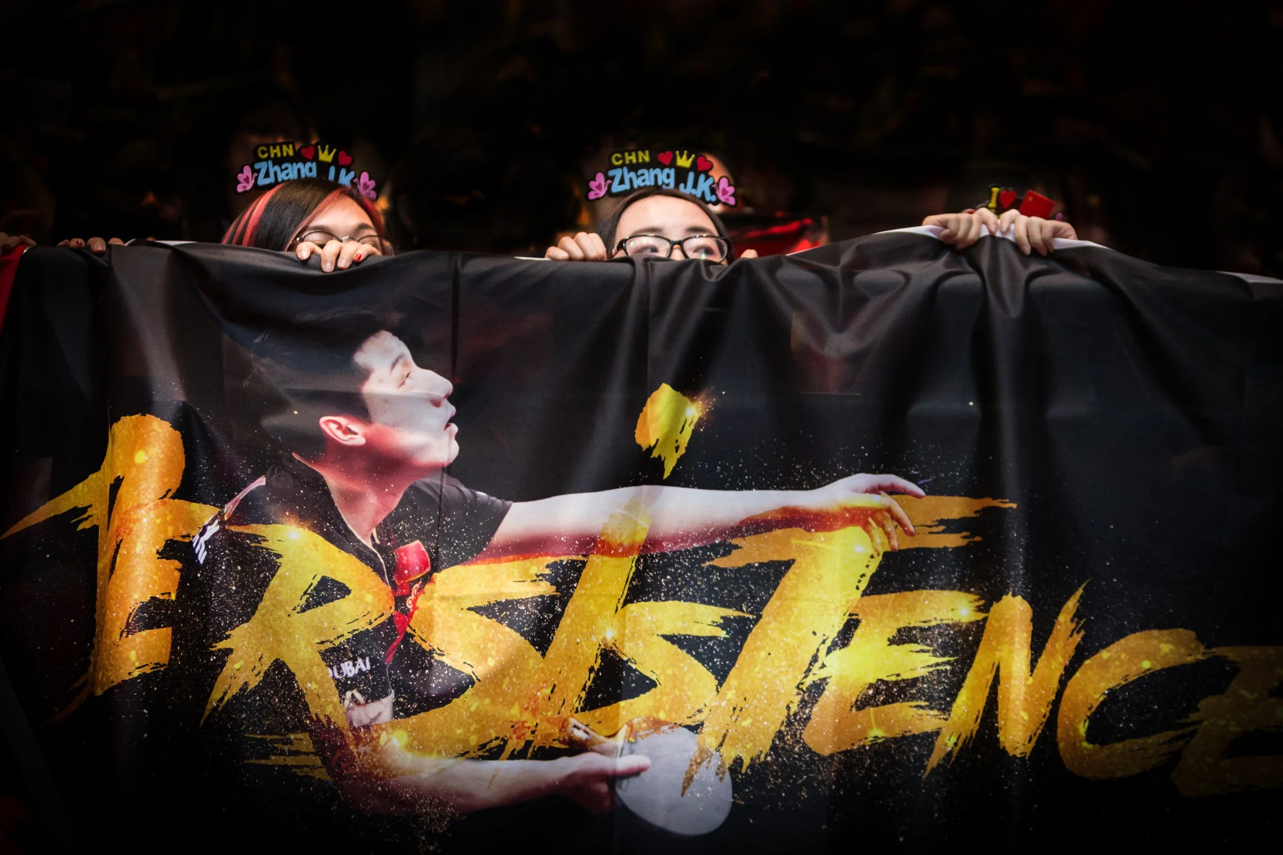 Four people wearing headbands with colorful decorations peek over a banner featuring a person playing esports or gaming.