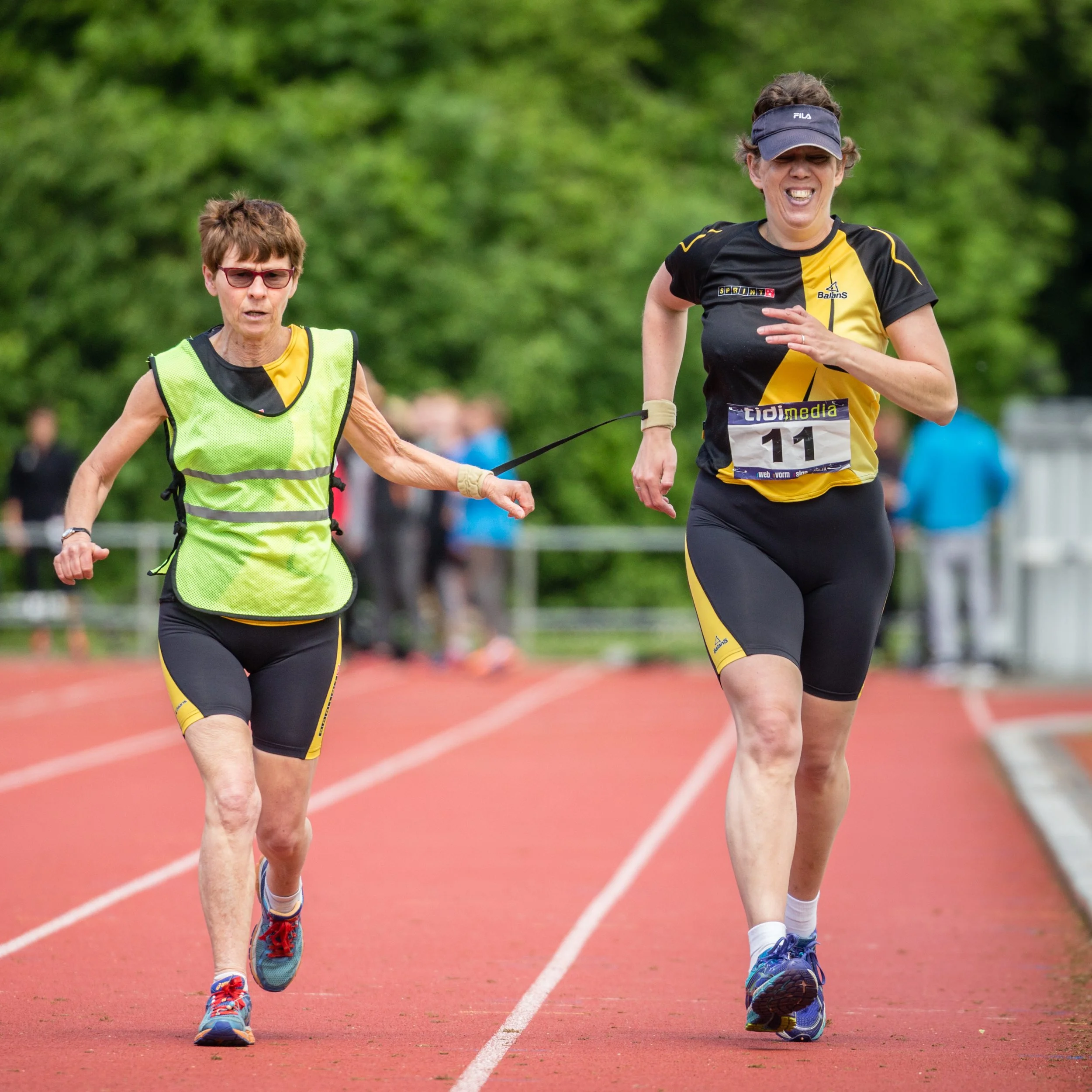Two women running on a red outdoor track, side by side, in athletic gear with trees in the background.