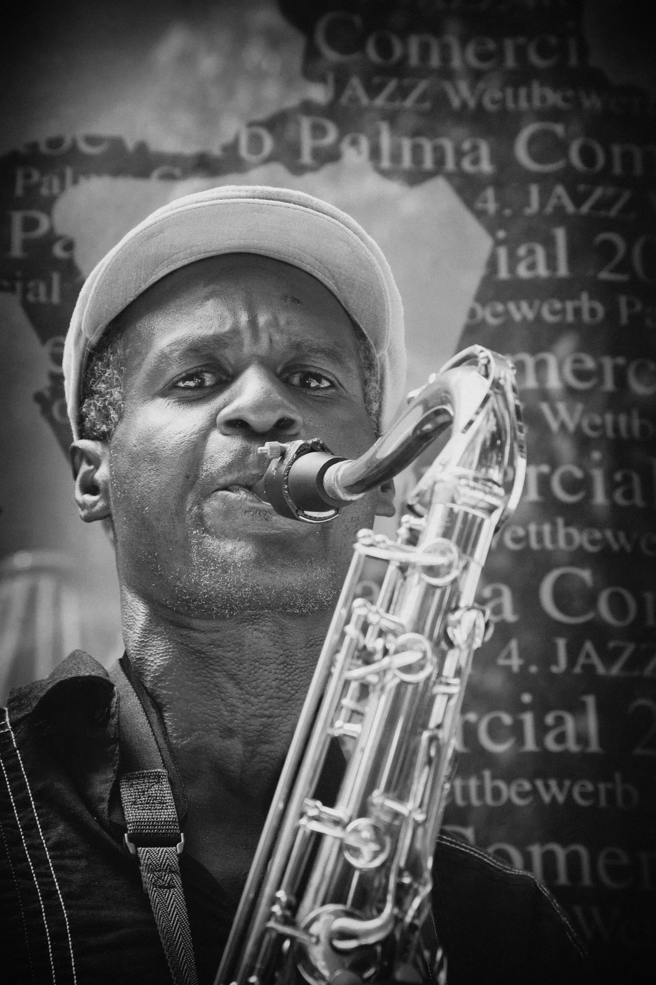 A black and white photo of a man playing a saxophone, wearing a hat and looking directly at the camera with a serious expression. In the background, there is text on a wall or banner that references jazz music.