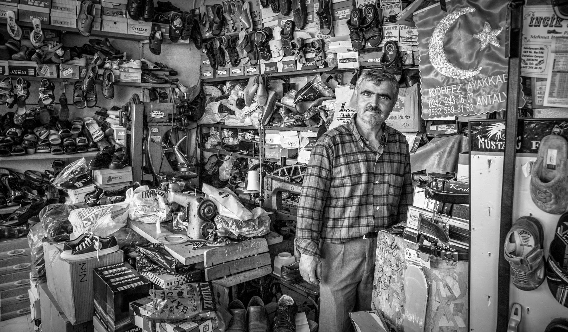 A man in a plaid shirt standing behind a counter in a shoe and accessory store filled with various shoes, boxes, and items. Shelves are lined with shoes and sandals, and there are boxes and plastic bags on the counter.
