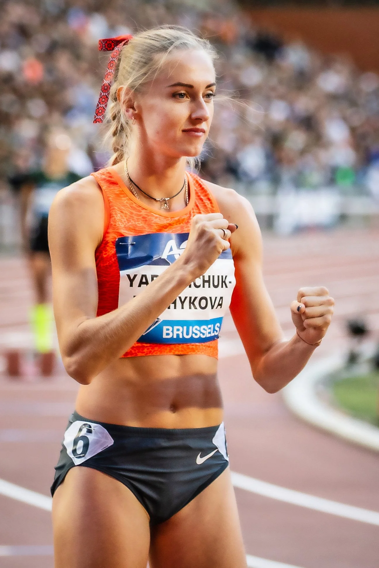A female athlete wearing a sleeveless orange top and black shorts appears to be running or warming up on a track during a race or event, with a blurred crowd in the background.