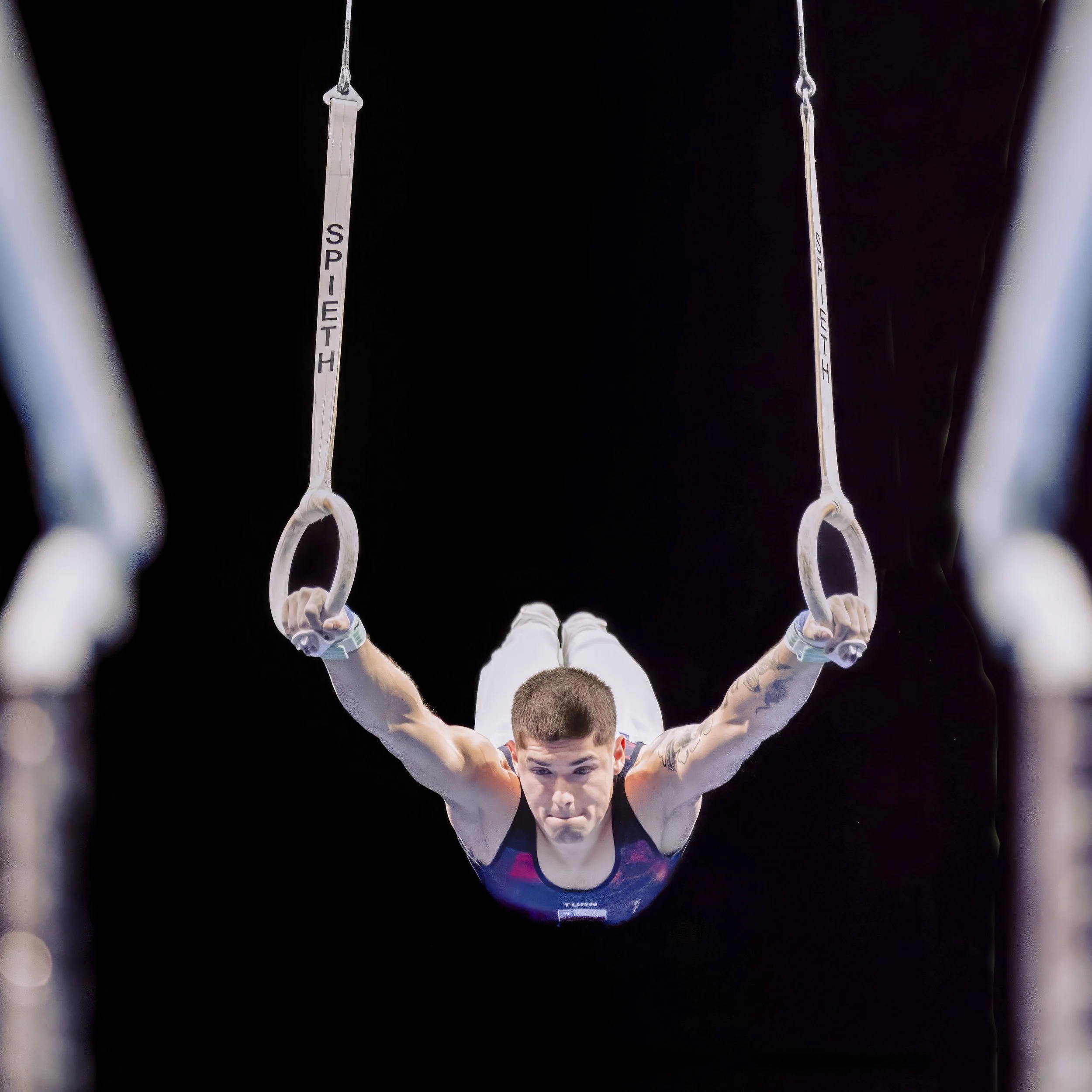 A male gymnast is performing a routine on the rings, holding his body straight and tense, with his face focused and intense, wearing a sports uniform with the name 'Turn' visible.