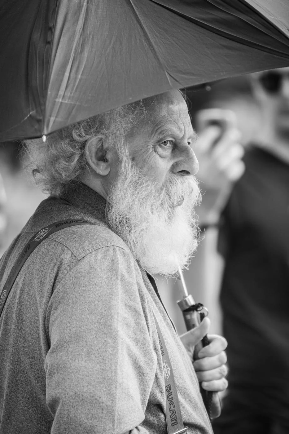 An elderly man with a white beard and curly hair holding an umbrella in front of him.
