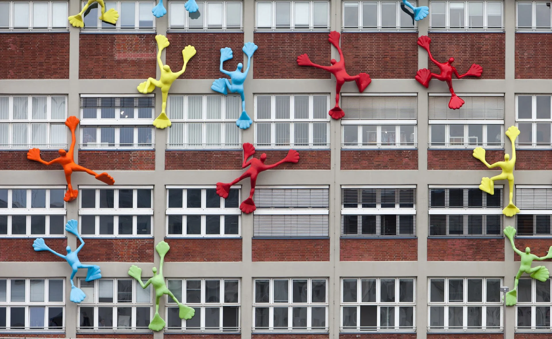 Colorful sculptures of climbing people in red, blue, yellow, orange, and green are mounted on the exterior of a multi-story brick building with numerous windows.