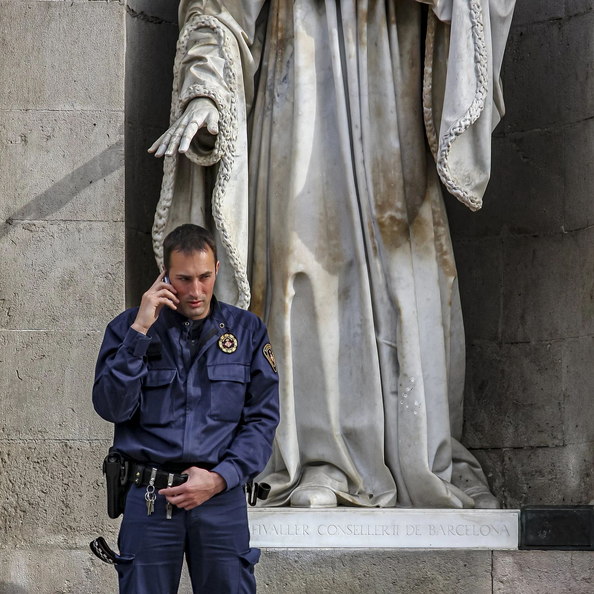 Police officer standing in front of a marble statue, talking on his cellphone, with a plaque reading 'FALLER CONSELLER DE BARCELONA' visible at the base of the statue.