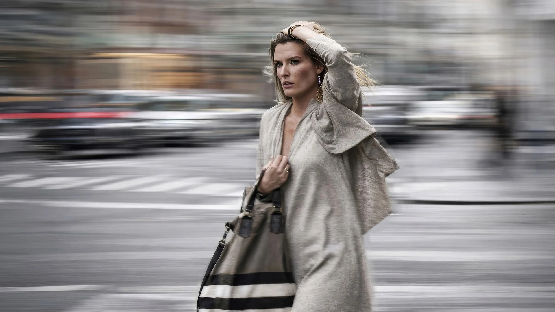 A woman walking through a city street with blurred cars in the background, wearing a light-colored trench coat and holding her hair back.