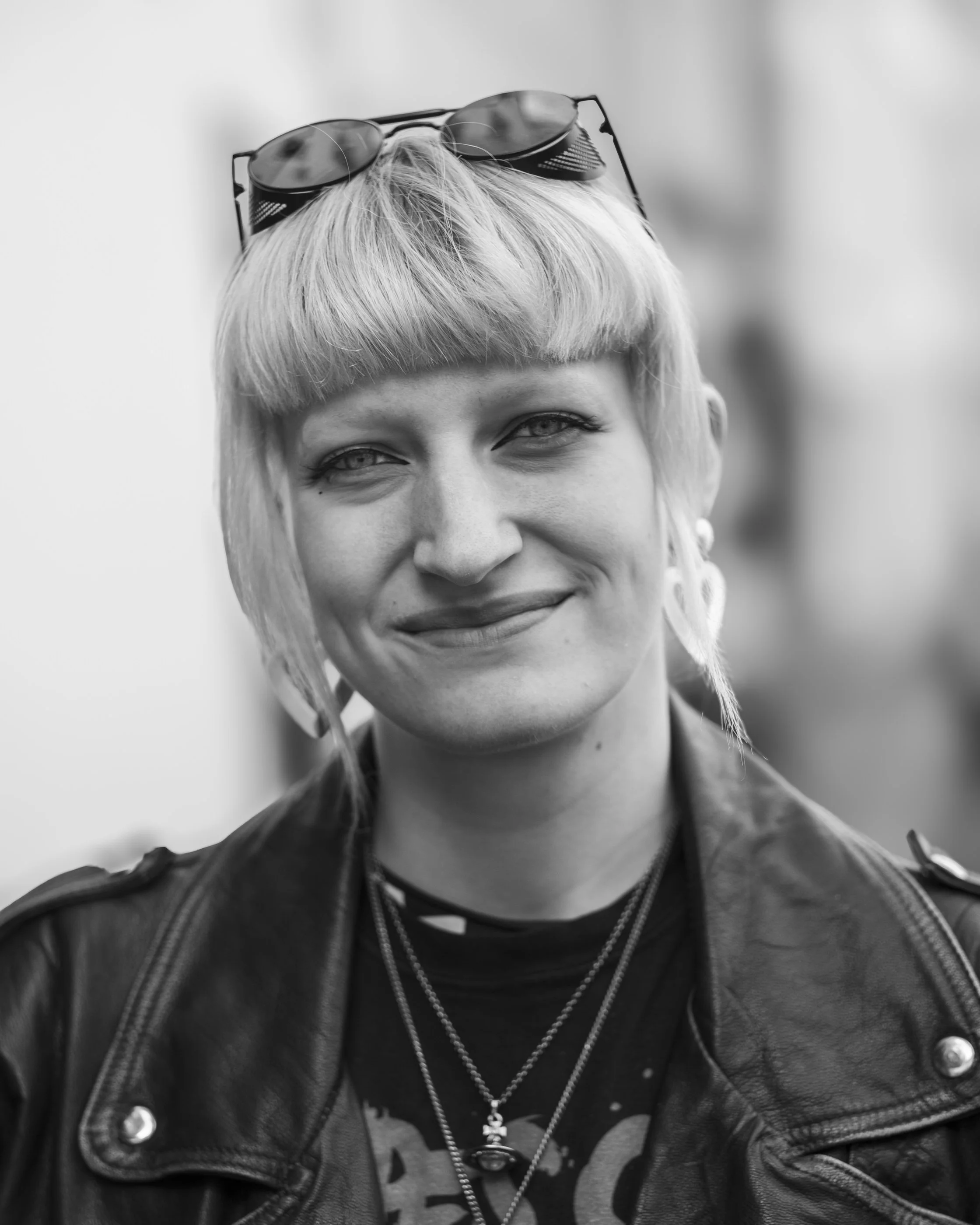 Close-up of a woman's face, with sunglasses on her head, short blonde hair, wearing a leather jacket and layered necklaces.