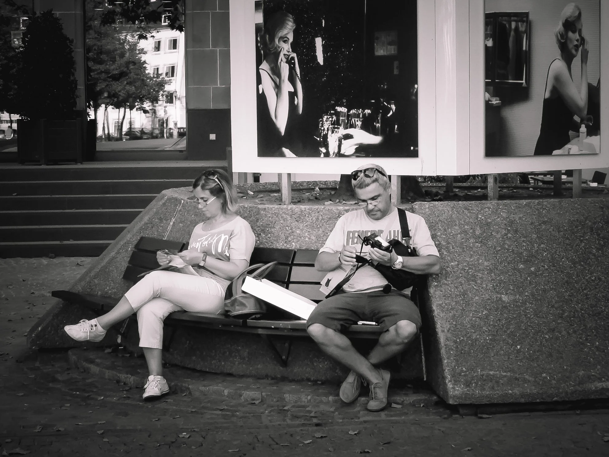 Two people sitting on a park bench, looking at their phones while a large billboard of a woman talking on a phone is visible behind them. The woman in the billboard is depicted in a glamorous style, and the scene is in black and white.