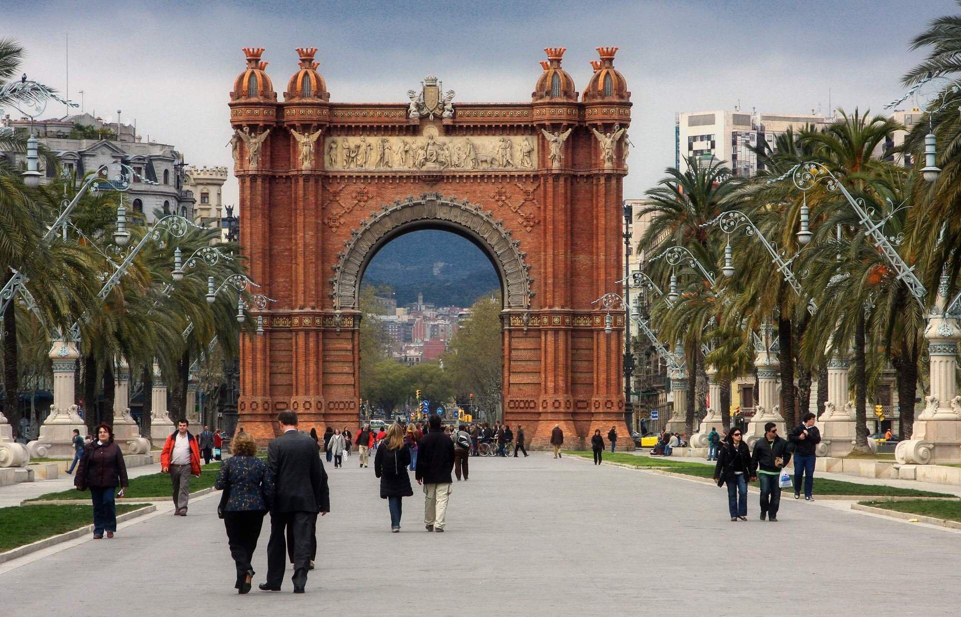 People walking through a street with tall palm trees lining both sides of the road, leading towards a large historic brick arch with ornate carvings and sculptures at the top, set against a cityscape with modern buildings and mountains in the backgro