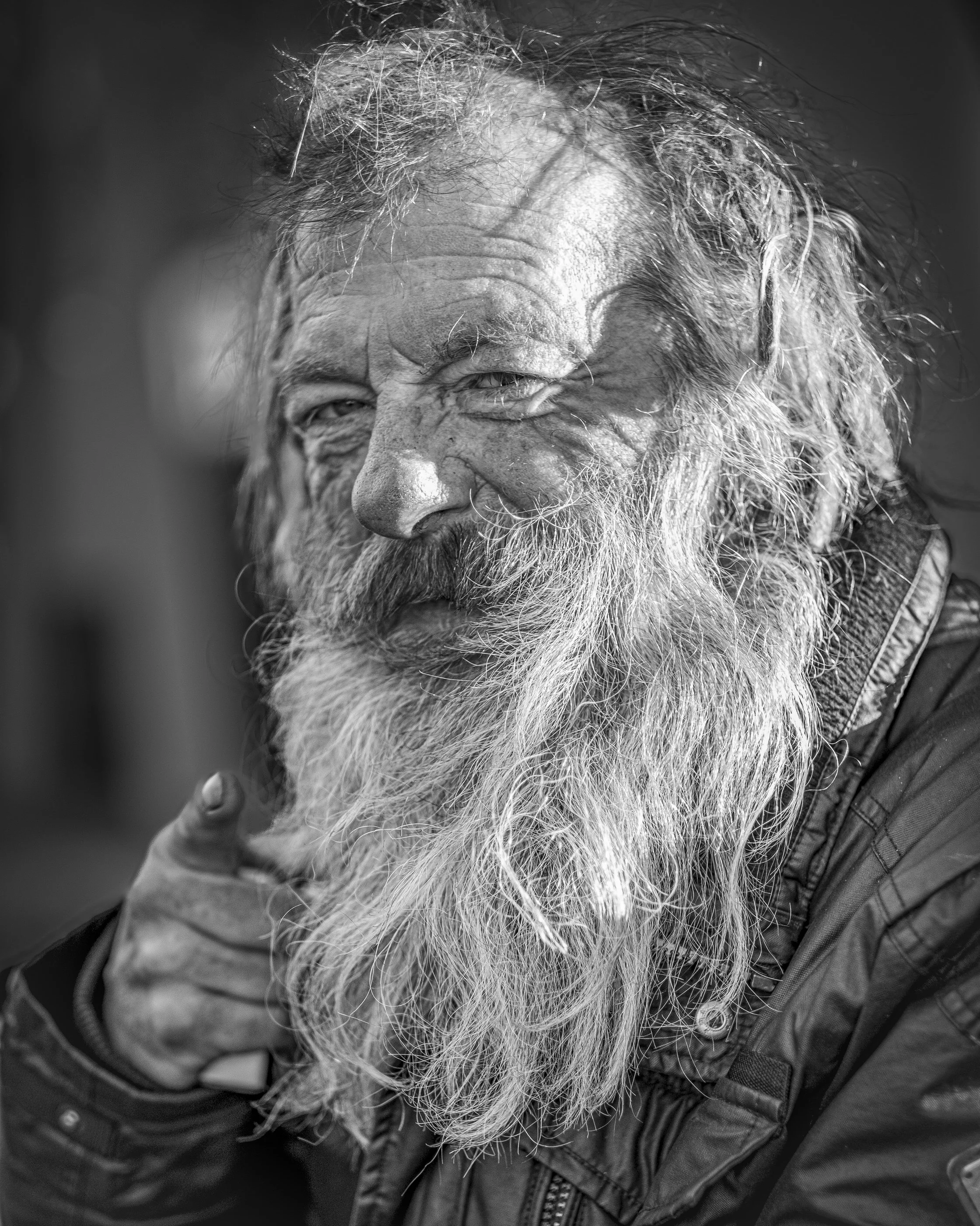 A close-up black and white photo of an elderly man with a full beard and disheveled hair, wearing a jacket.