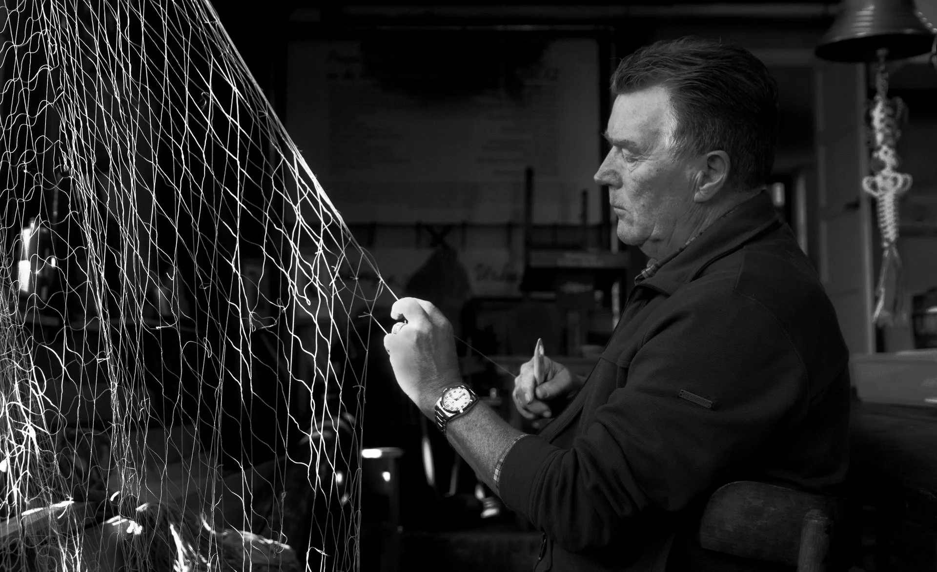 A man working with a large fishing net in a workshop or garage, with shelves and tools visible in the background.