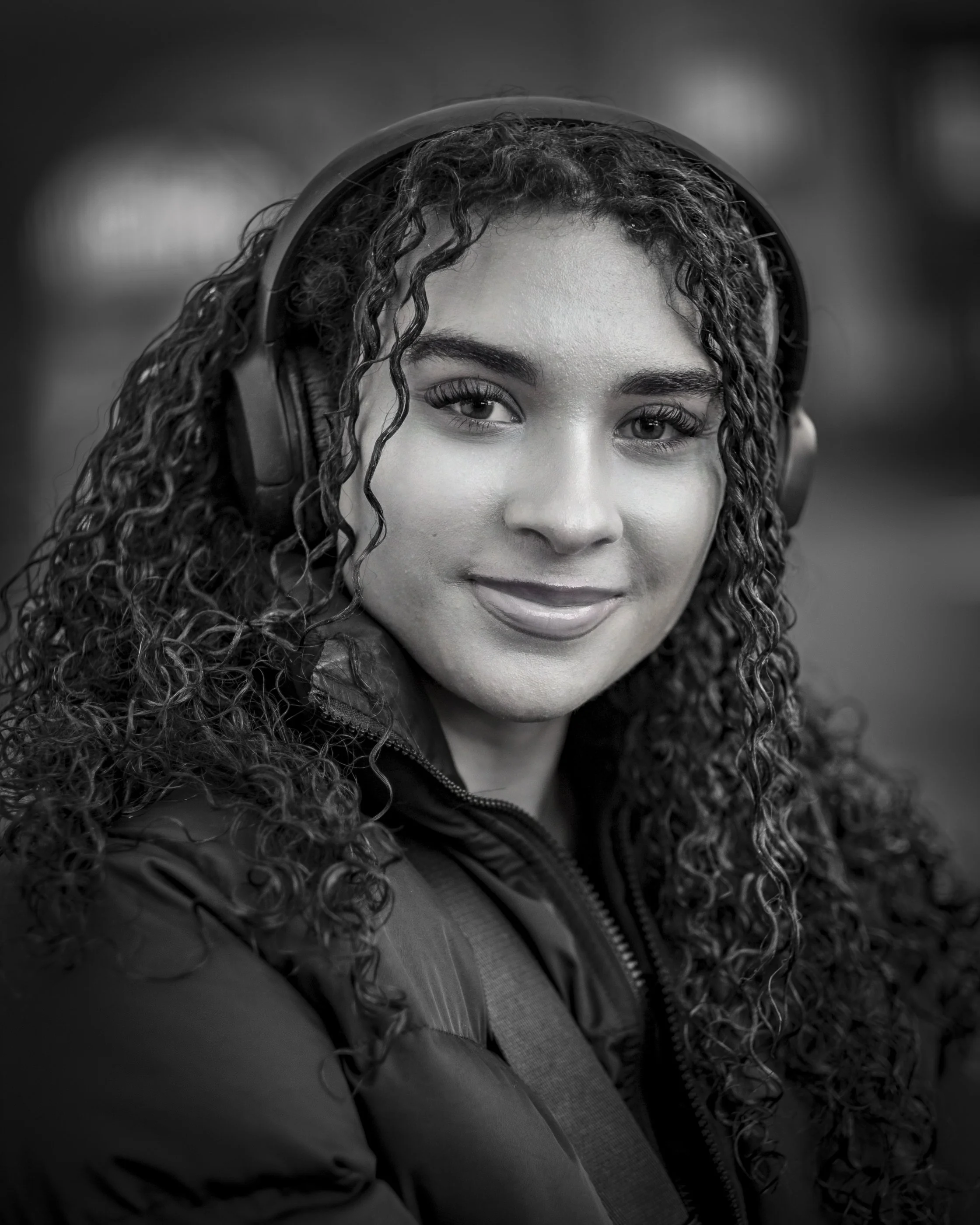 A close-up, black-and-white photograph of a young woman with curly hair wearing headphones and smiling softly at the camera.