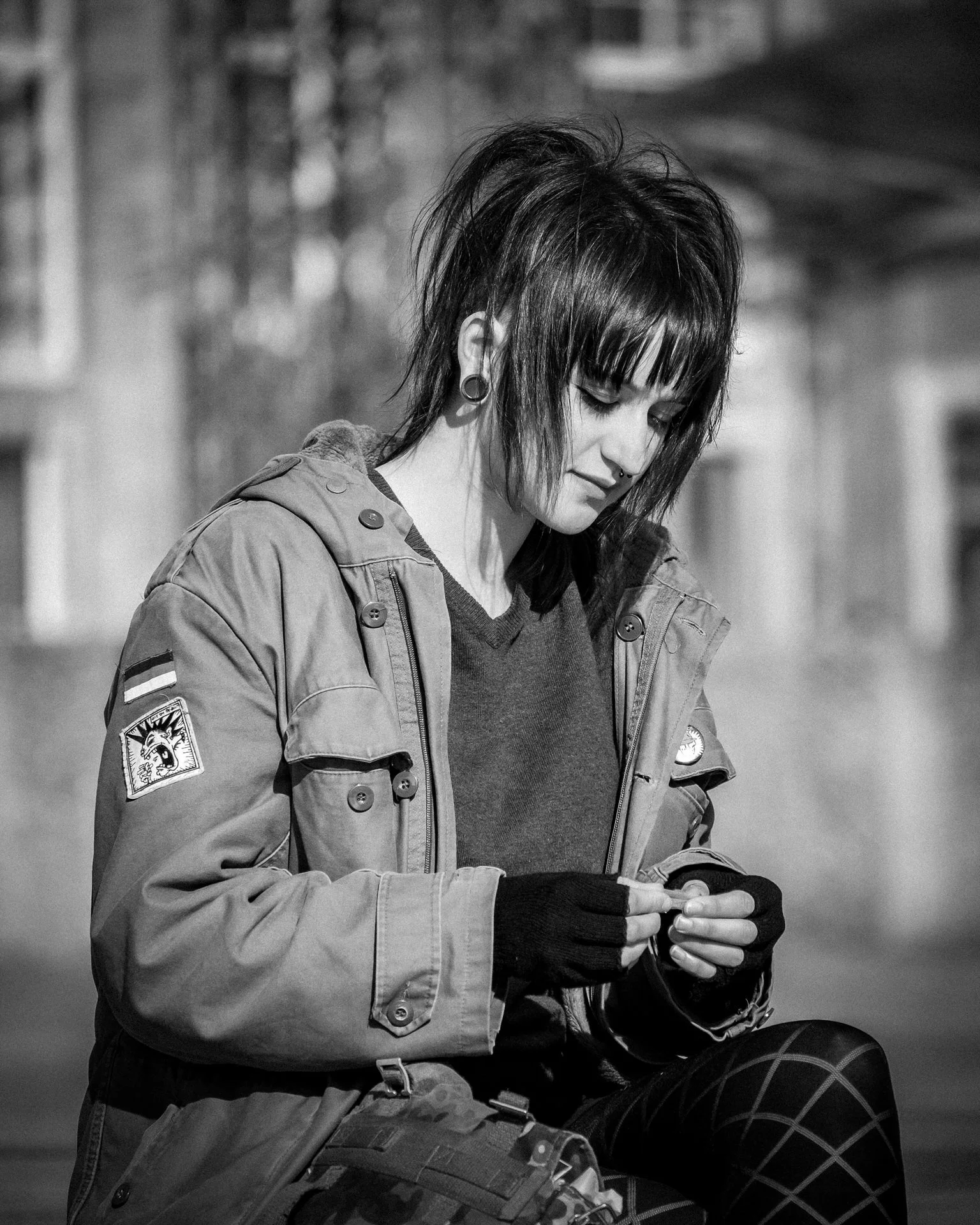 A young woman with dark, spiky hair and piercings, wearing a jacket with patches, looking down at her phone in an outdoor setting.