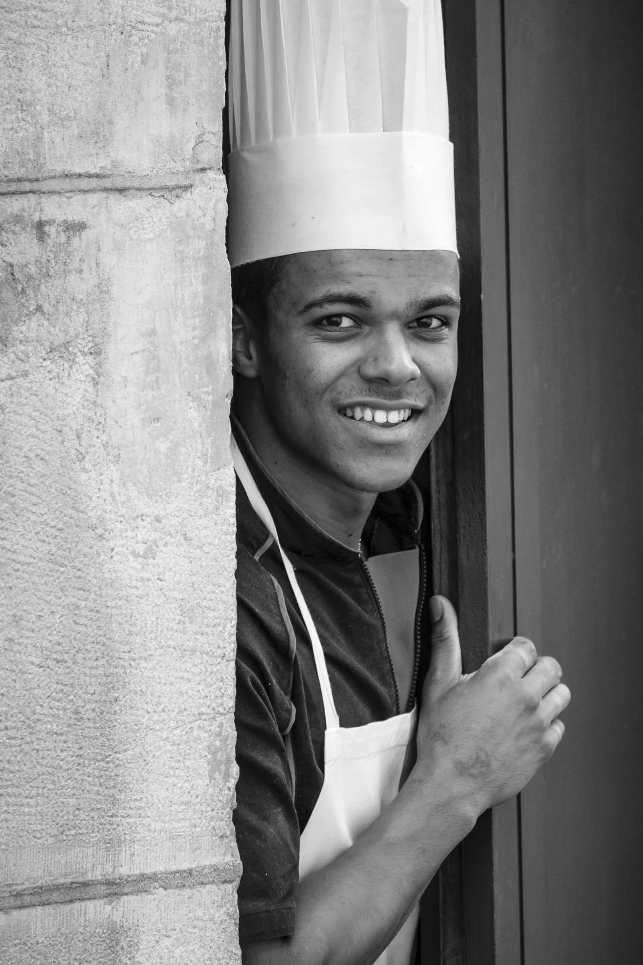 Young man wearing a chef's hat and apron smiling and giving a thumbs-up while peeking from behind a wall.