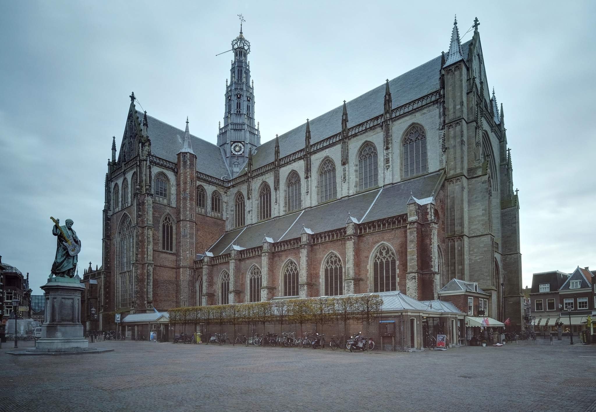 A large Gothic-style church with tall spires and stained glass windows. A statue of a person holding a cross stands in the foreground. Bicycles and scooters are parked at the base of the church on a paved square.