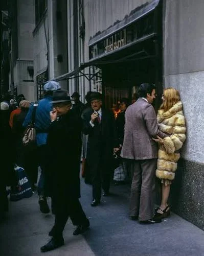 A man and woman talking outside a shop on a city street, with several pedestrians walking by, some looking at their phones or carrying bags.