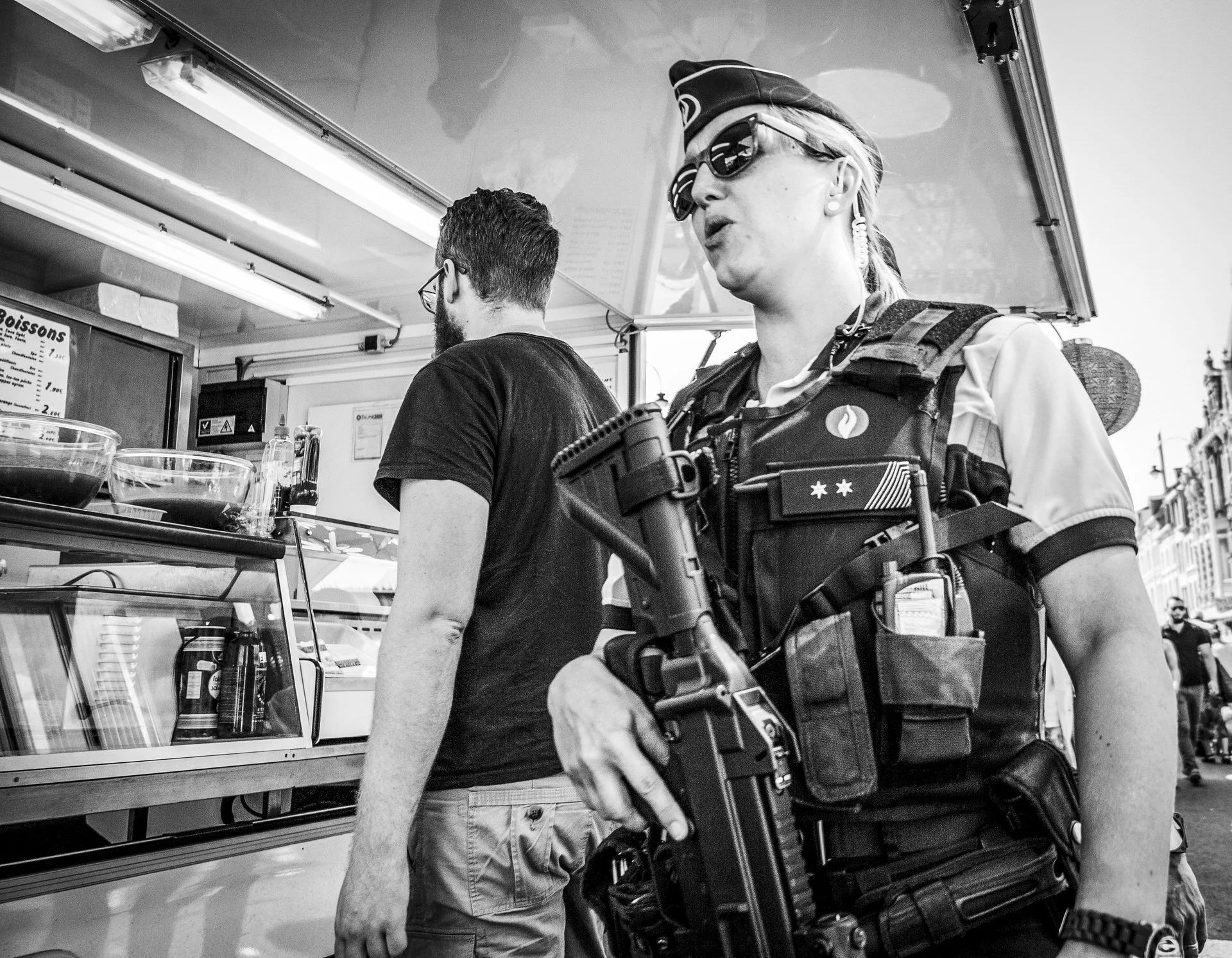 A woman in sunglasses and a hat, wearing a tactical vest, holding a handgun, standing on a city street with pedestrians and buildings in the background.