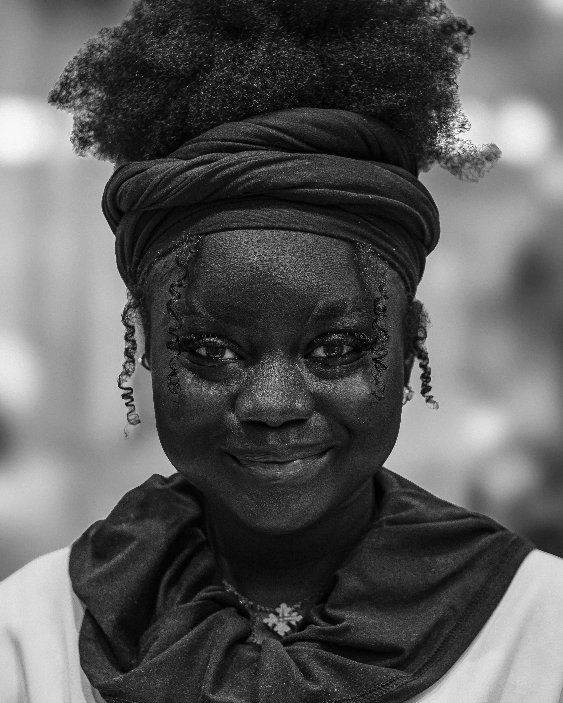 A black and white photo of a woman with dark skin and curly hair, wearing a head scarf, looking directly at the camera with a slight smile.