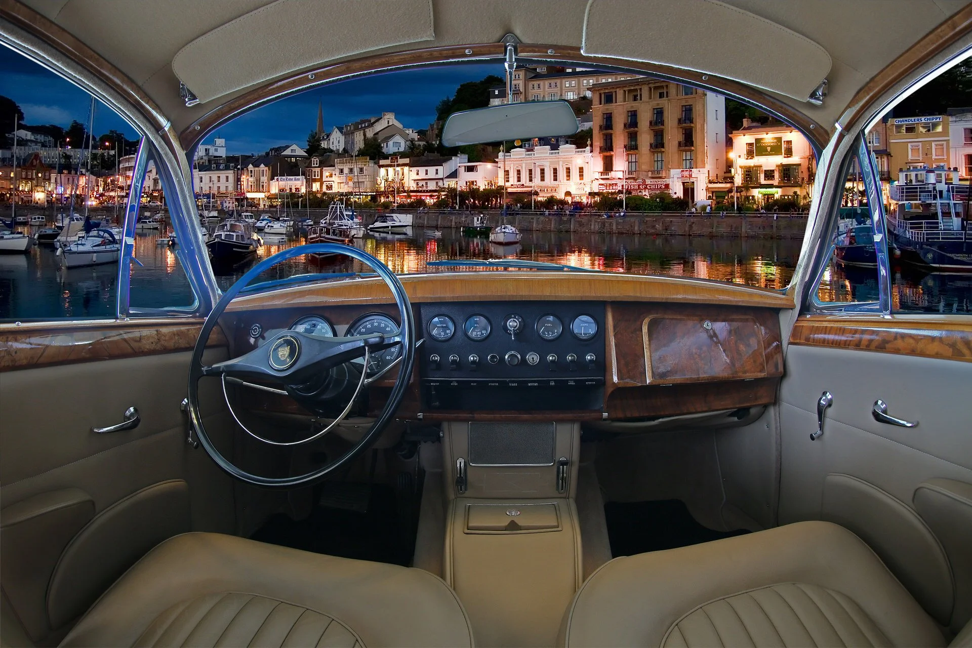 View from inside a vintage car looking out onto a harbor at dusk, with boats docked and a town skyline with buildings and lights in the background.