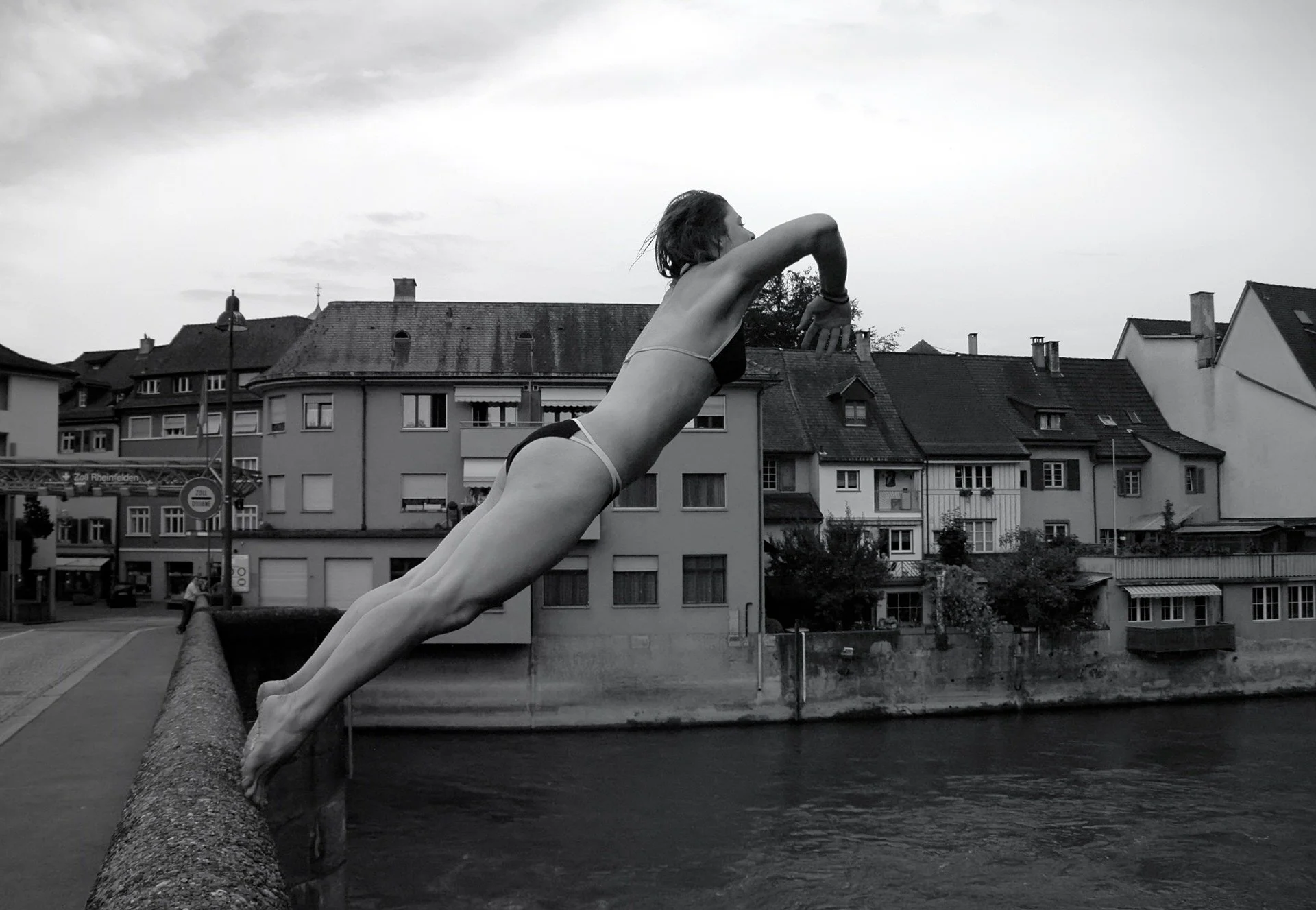 A black and white photograph of a large sculpture of a woman stretching forward, lying parallel to a stone wall with her left arm extended. There is a row of residential buildings with pitched roofs in the background under an overcast sky.