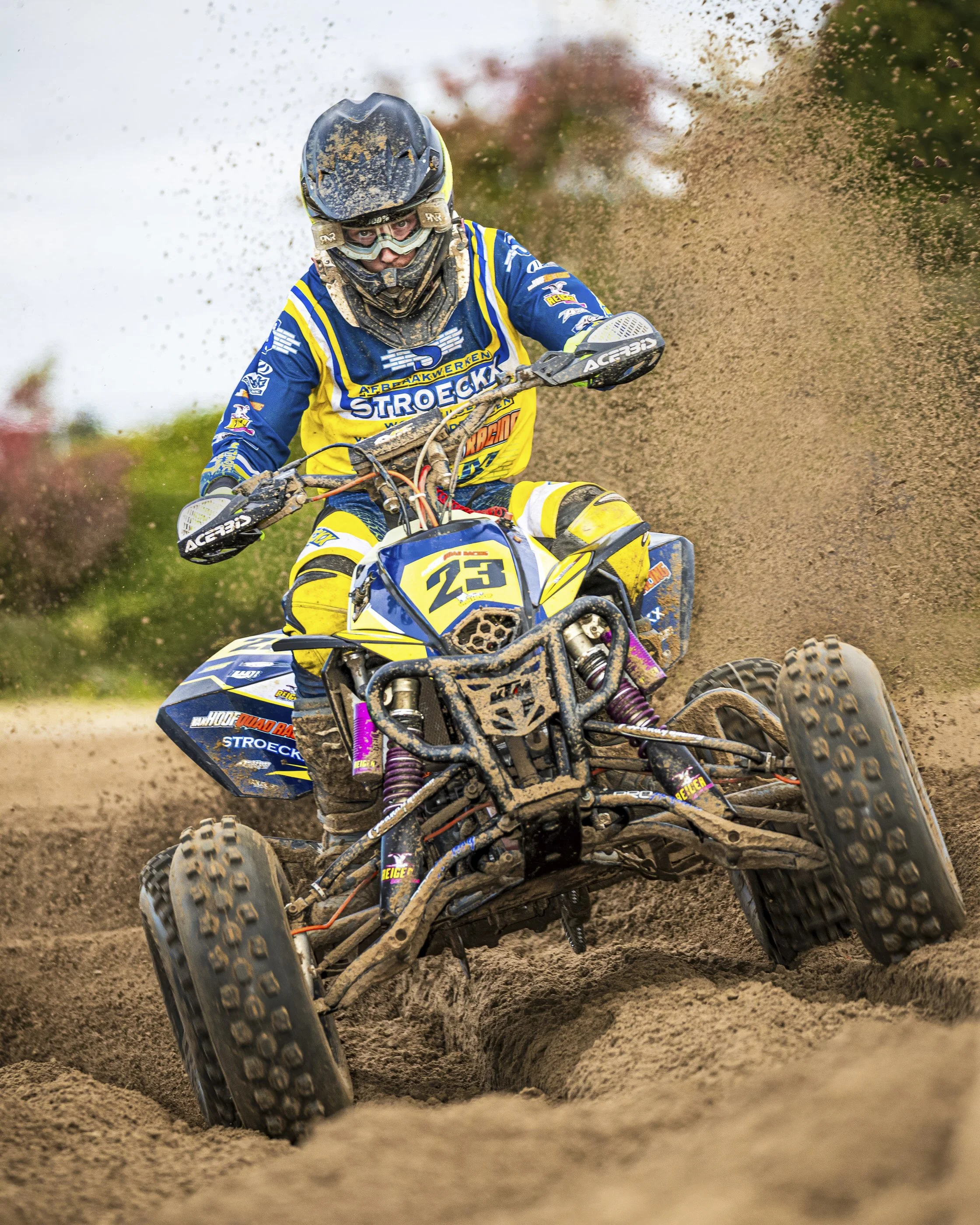 A man wearing a helmet and goggles riding a dirt bike through a muddy off-road track. The rider is dressed in blue and yellow gear and is kicking up dirt as he navigates the terrain.