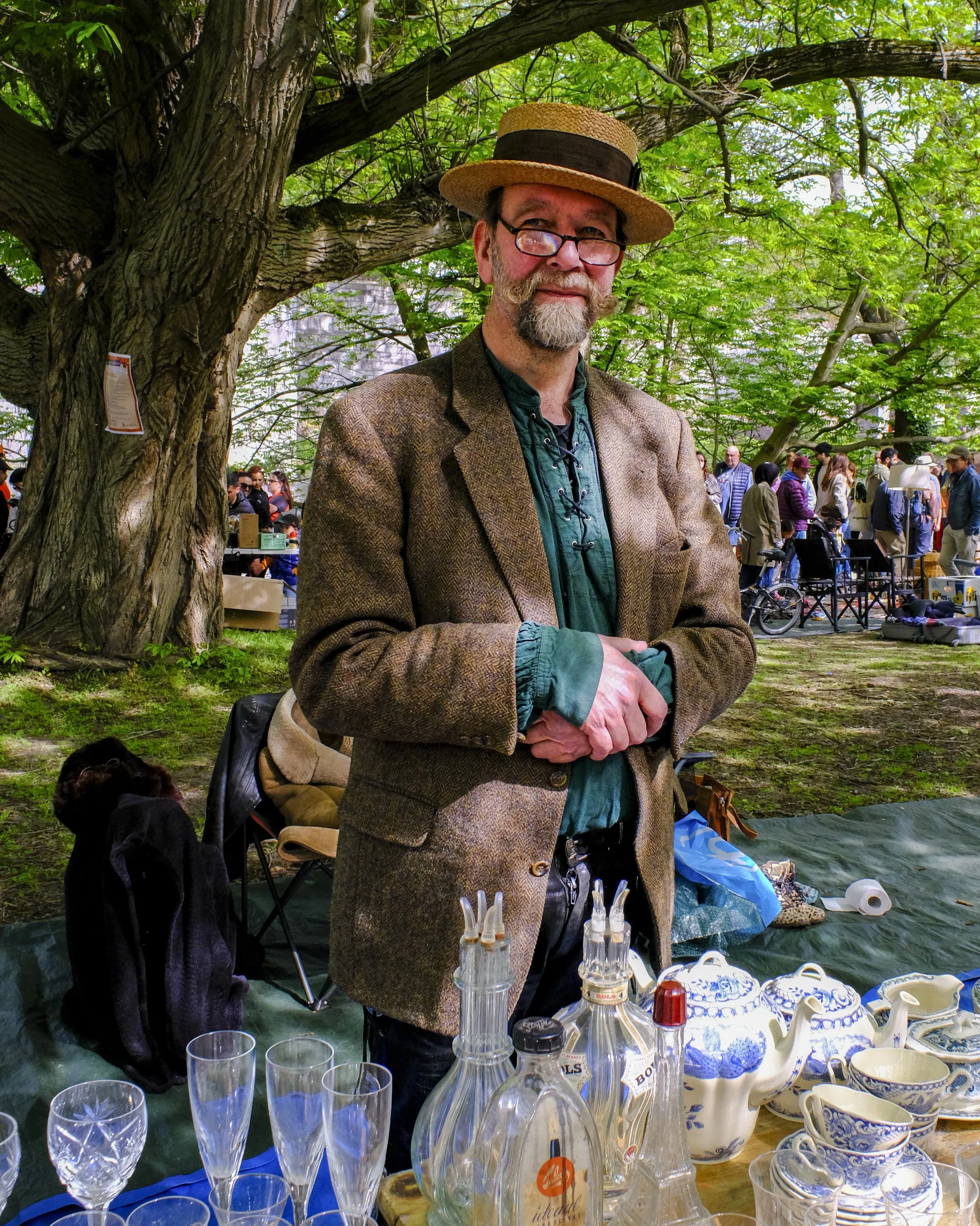 A man with a beard and glasses wearing a straw hat and a tweed jacket is standing outdoors in front of a large tree, with a crowd in the background. The scene appears to be a market or fair with various glassware, porcelain, and bottles displayed on 